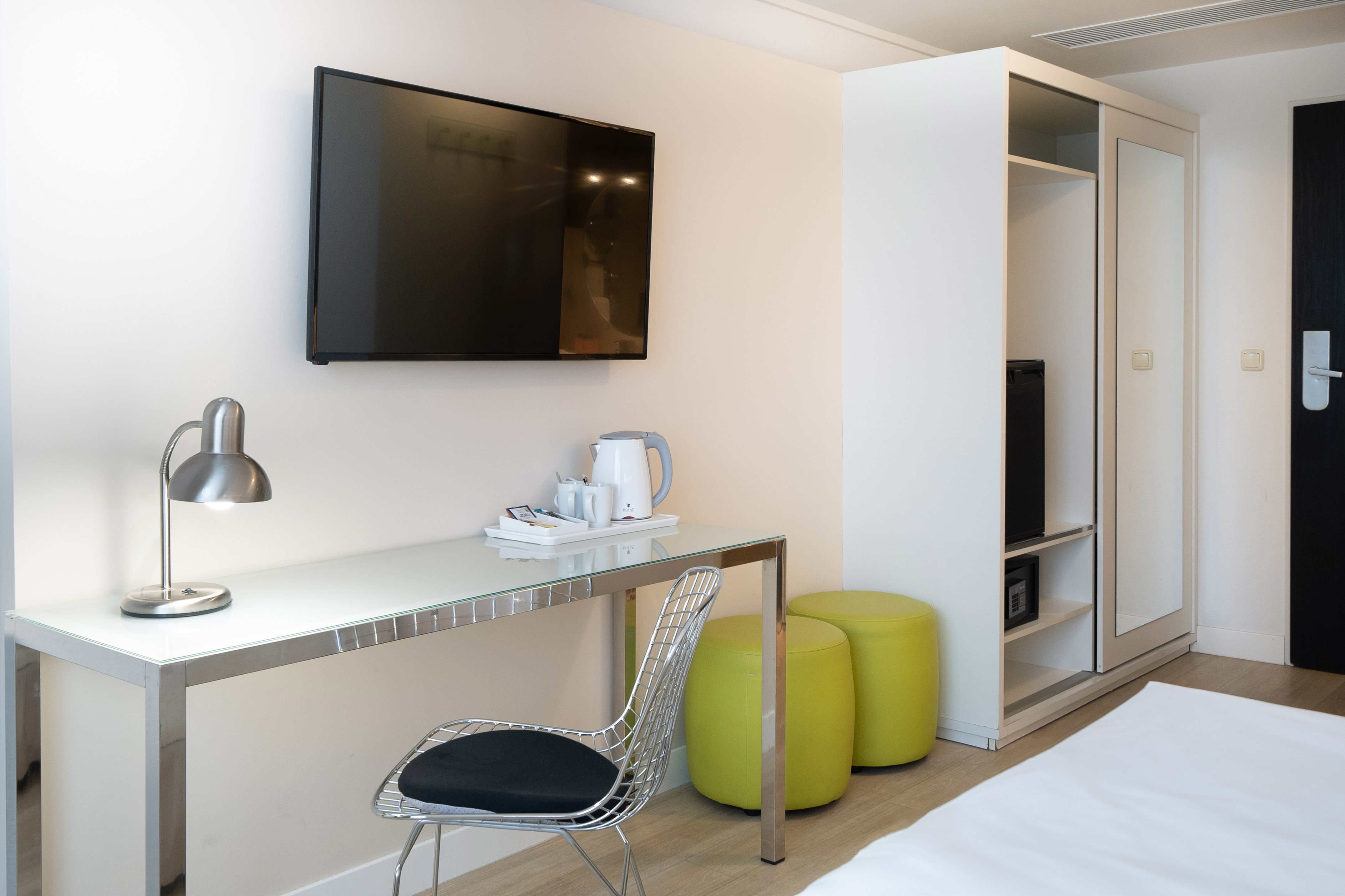 Modern hotel room with wall TV, glass desk, kettle, and lime green ottomans.