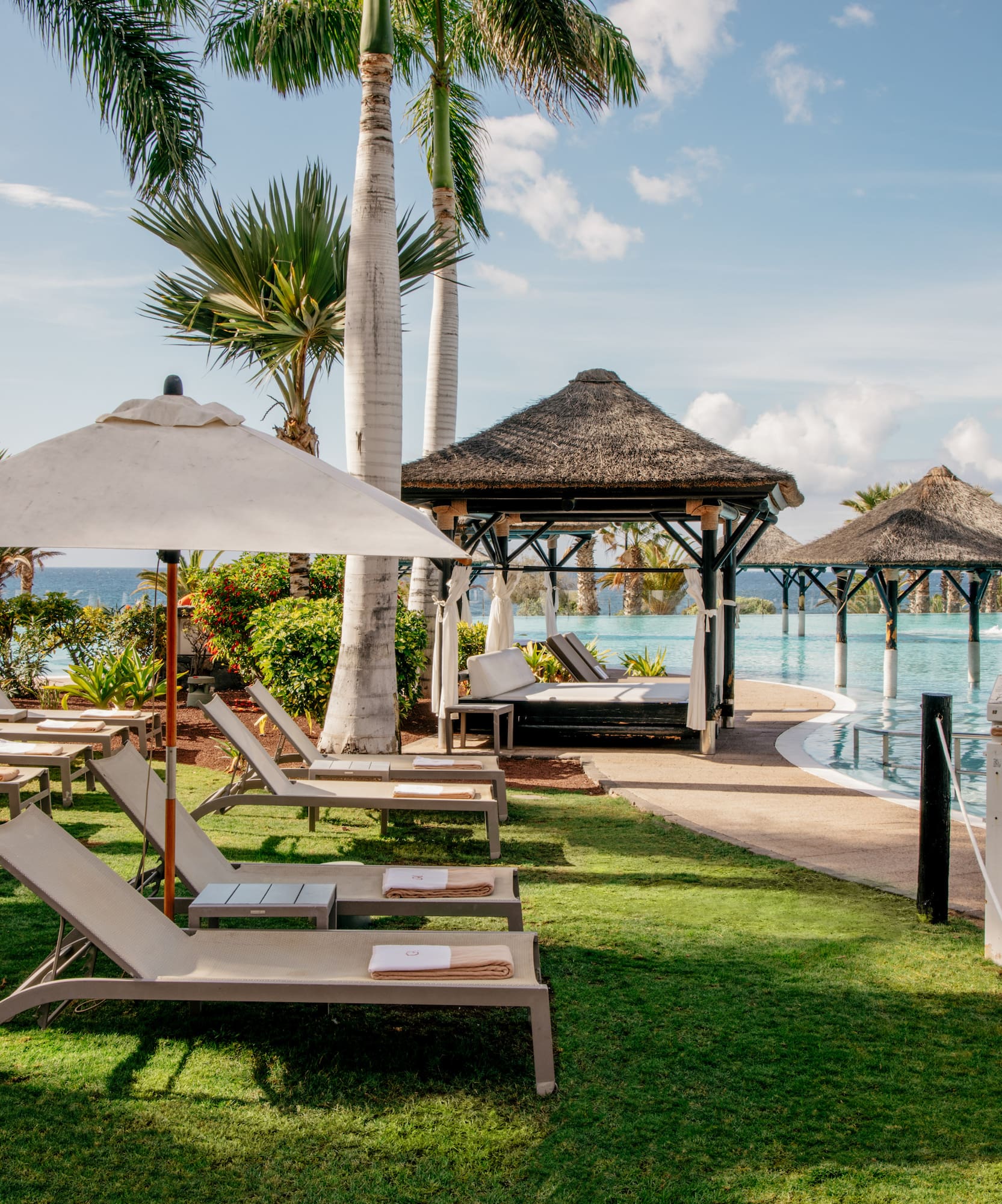 Tropical resort pool area with lounge chairs, palm trees, and thatched gazebos.