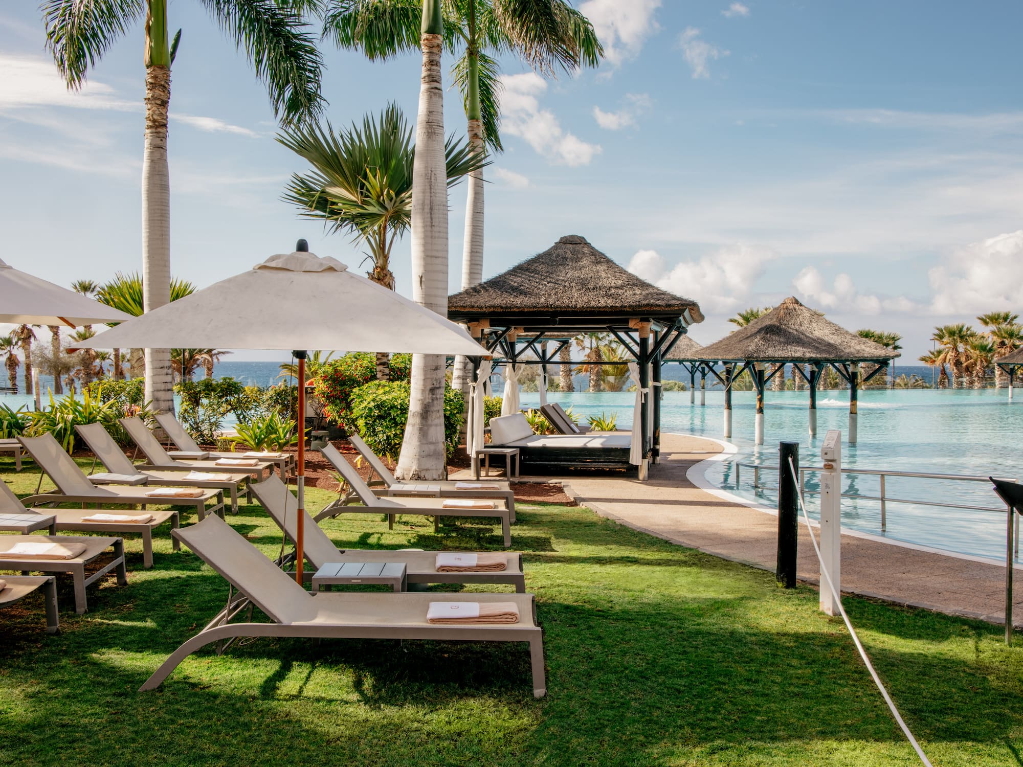 Tropical resort pool area with lounge chairs, palm trees, and thatched gazebos.