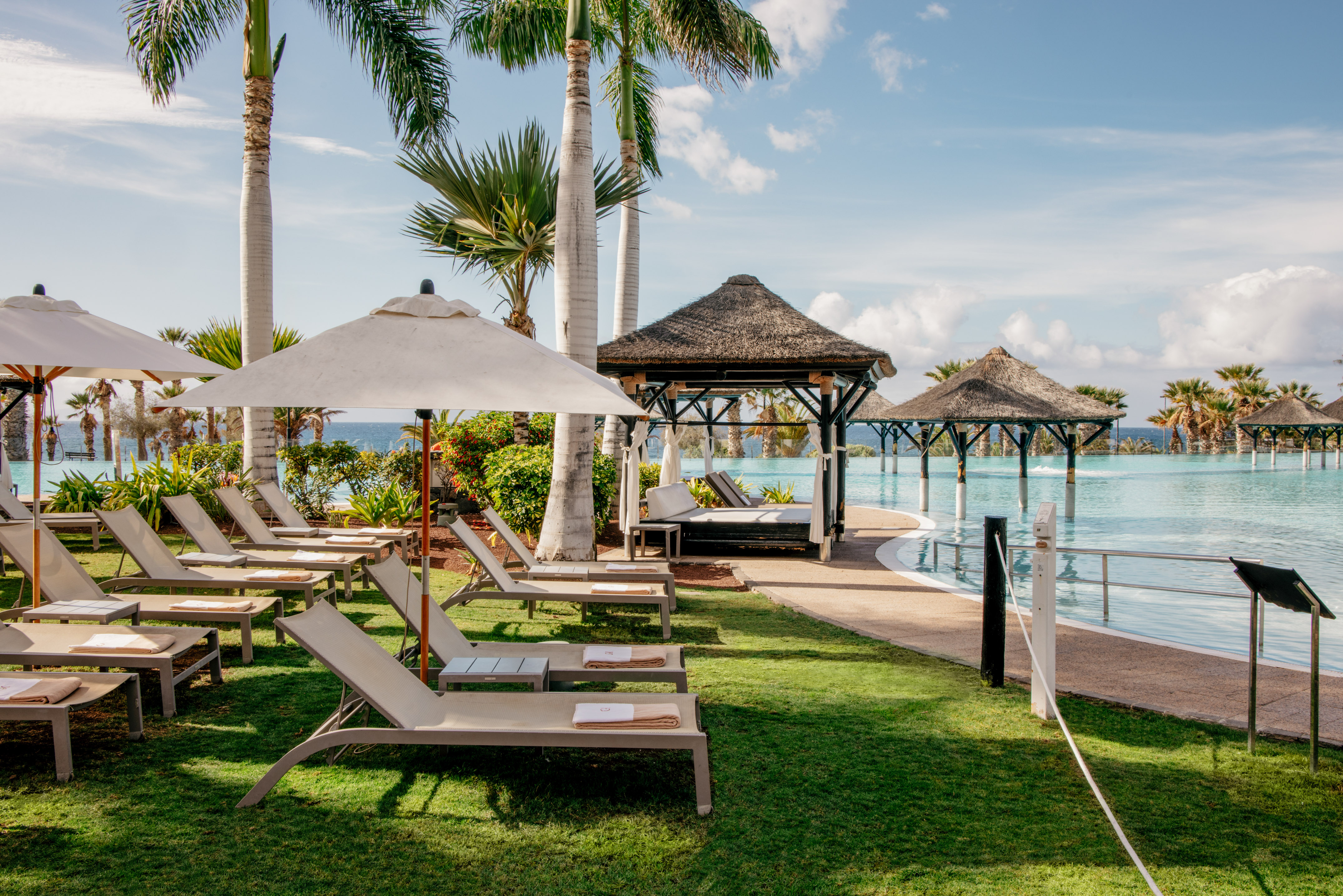 Tropical resort pool area with lounge chairs, palm trees, and thatched gazebos.