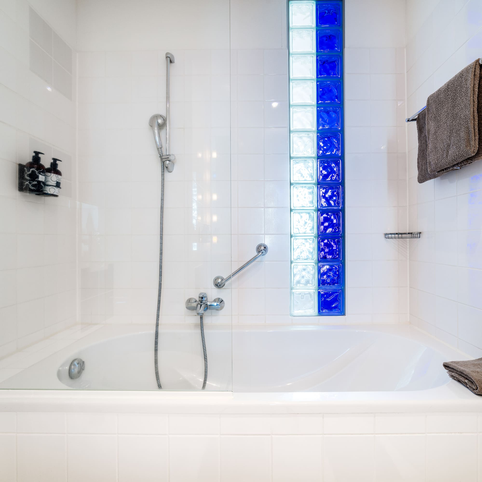 Bathtub and shower in a white tiled bathroom with blue glass blocks.