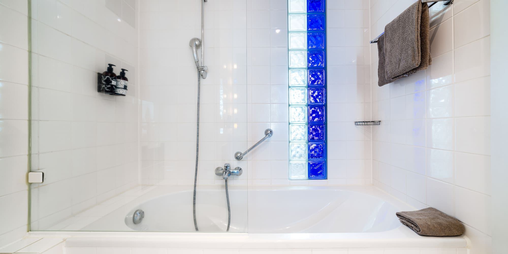 Bathtub and shower in a white tiled bathroom with blue glass blocks.