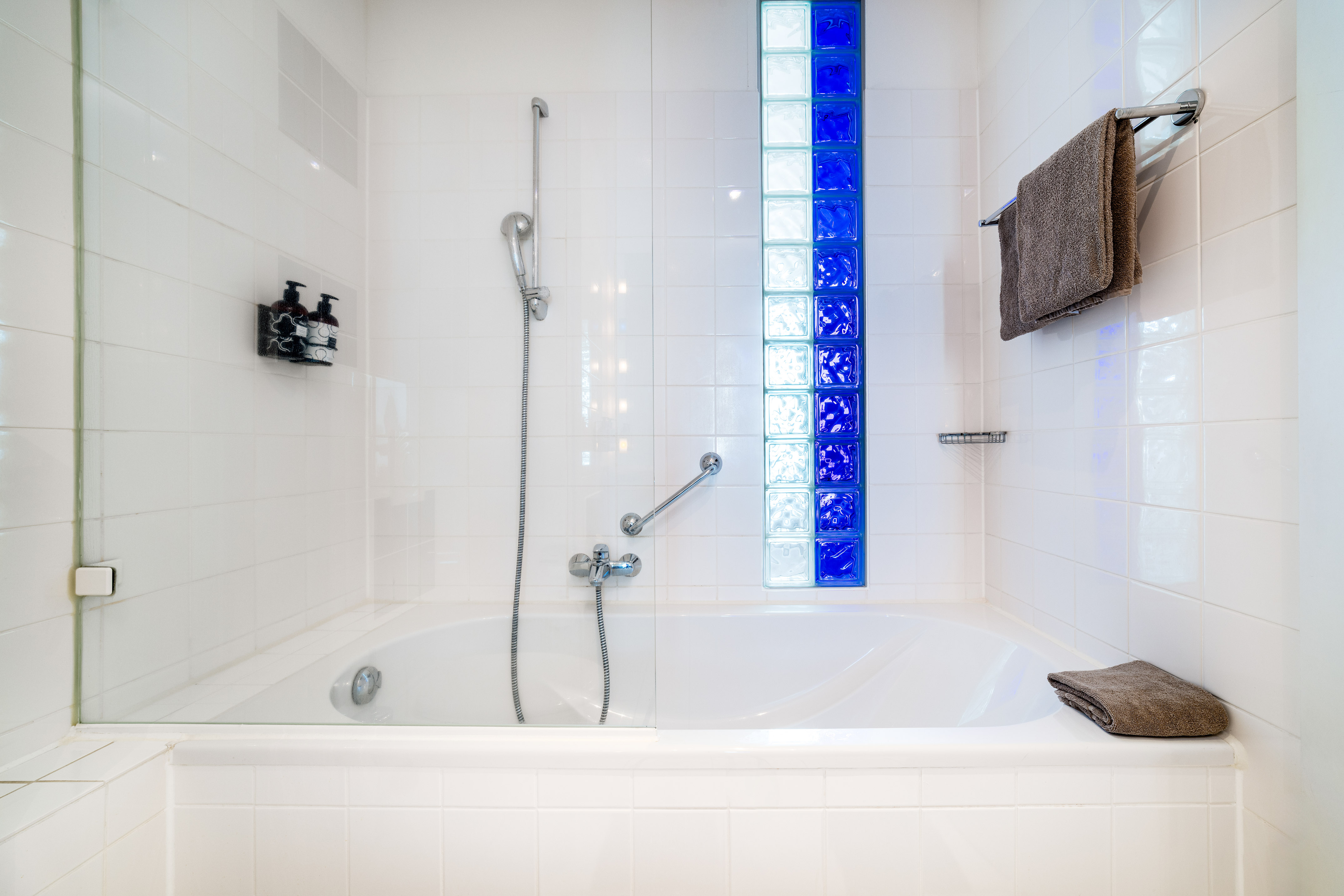 Bathtub and shower in a white tiled bathroom with blue glass blocks.