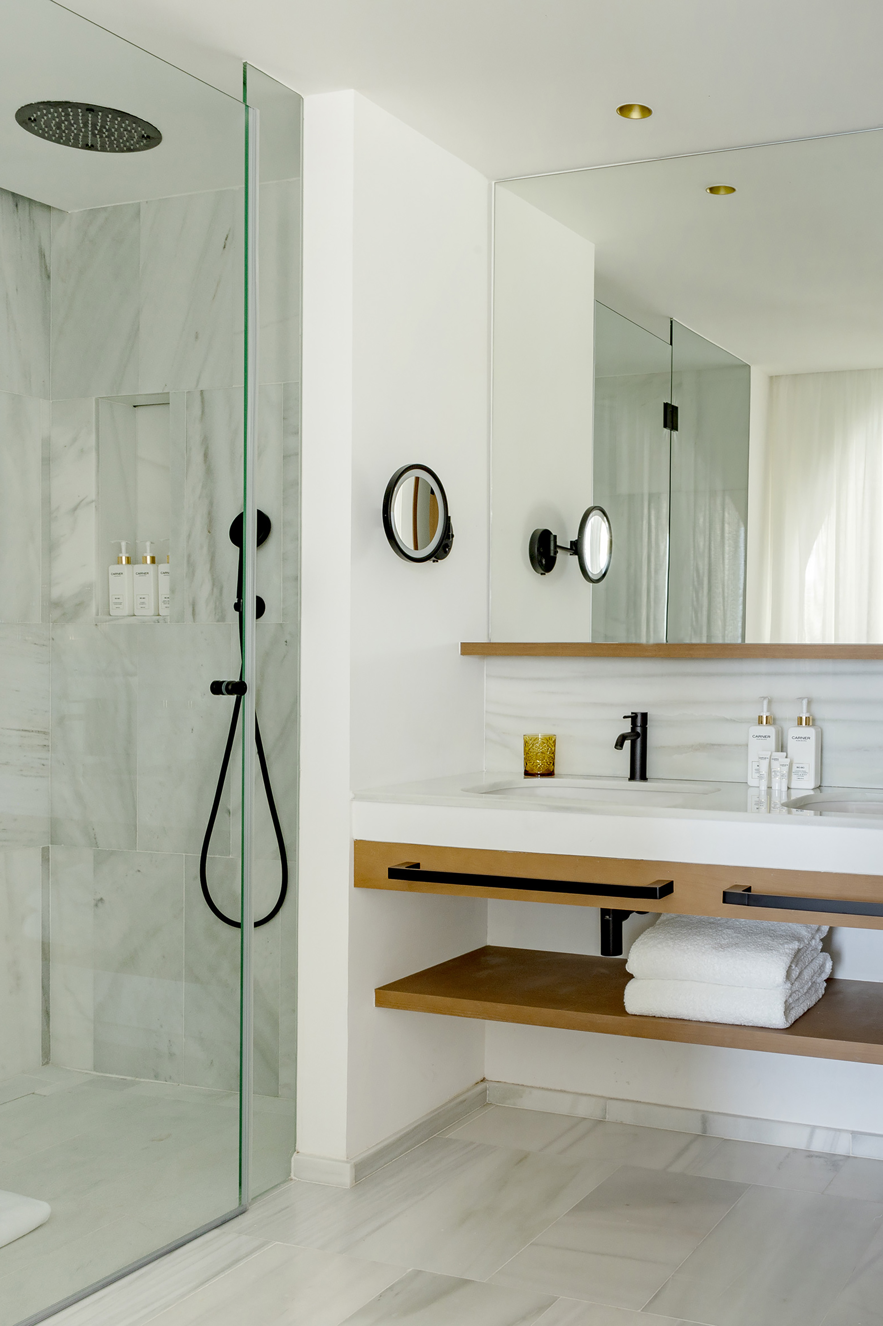 Luxury hotel bathroom with marble shower, black fixtures, white vanity, and wood shelf.