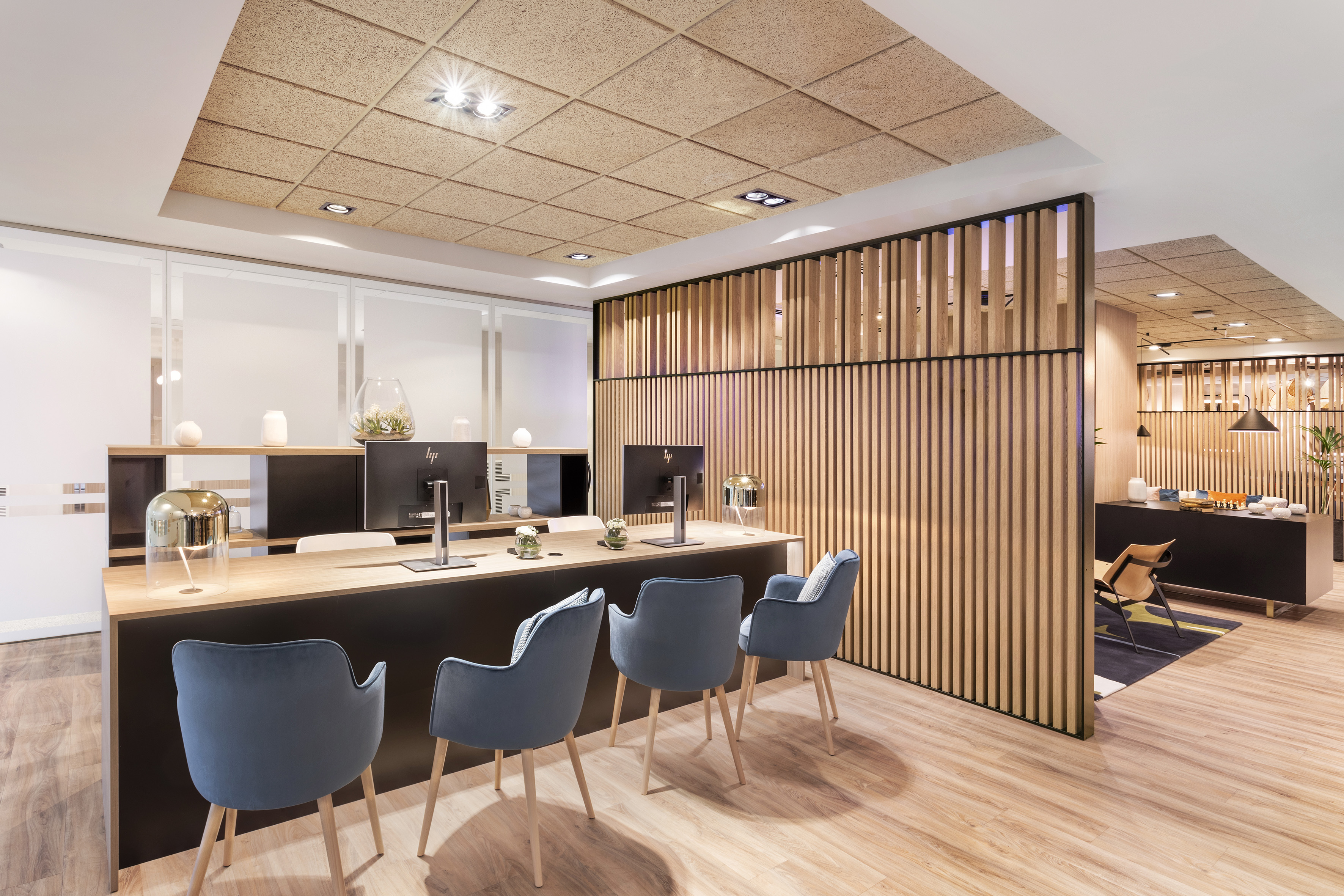 Modern office reception desk with wood slat partition, acoustic ceiling, and blue chairs.