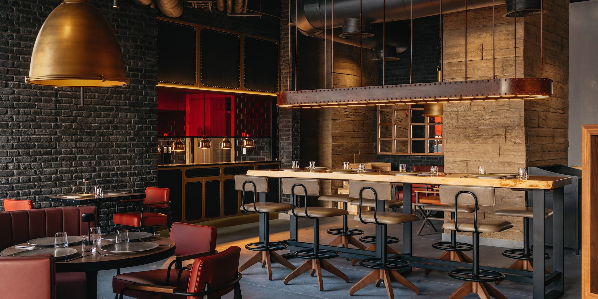 Industrial restaurant interior: dark brick, brass lights, communal bar, and red seating.