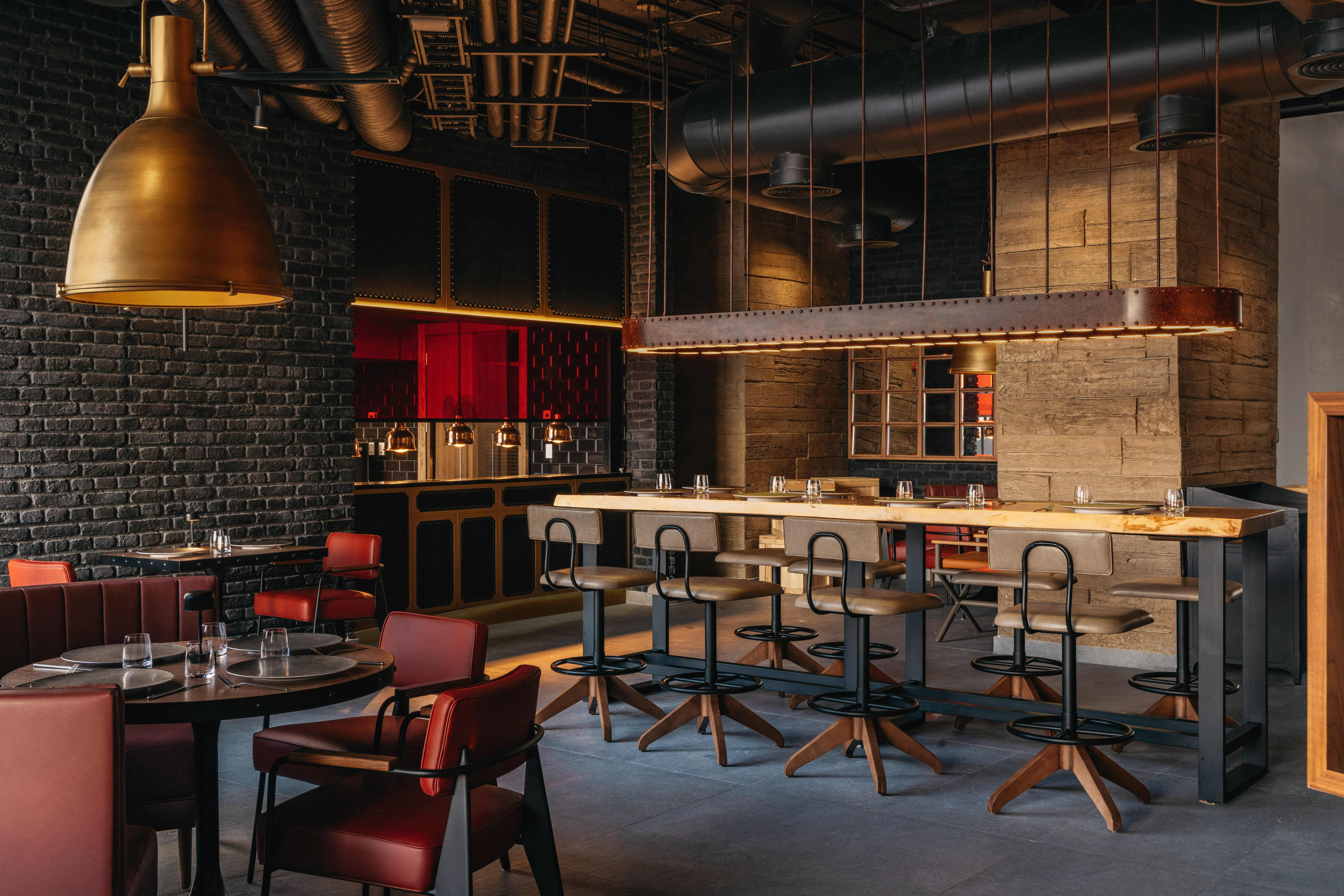Industrial restaurant interior: dark brick, brass lights, communal bar, and red seating.