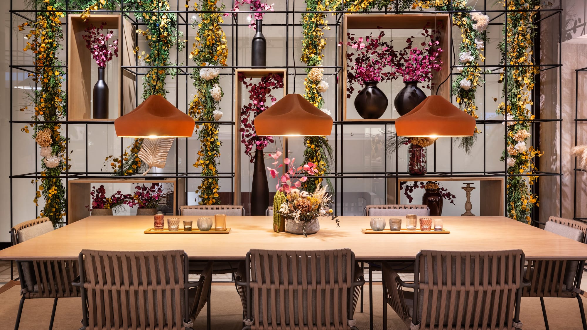 Modern dining room: wooden table, woven chairs, orange pendant lights, floral decor.