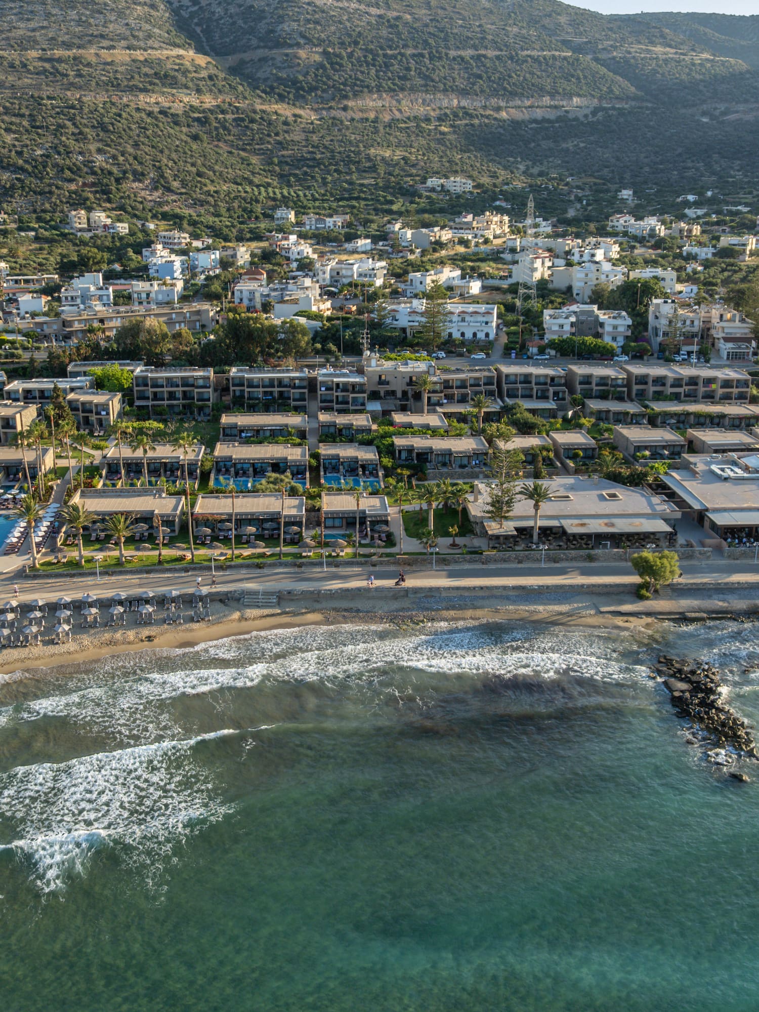 Aerial view: Beachfront resort with pools, sandy beach, clear ocean, and mountains.