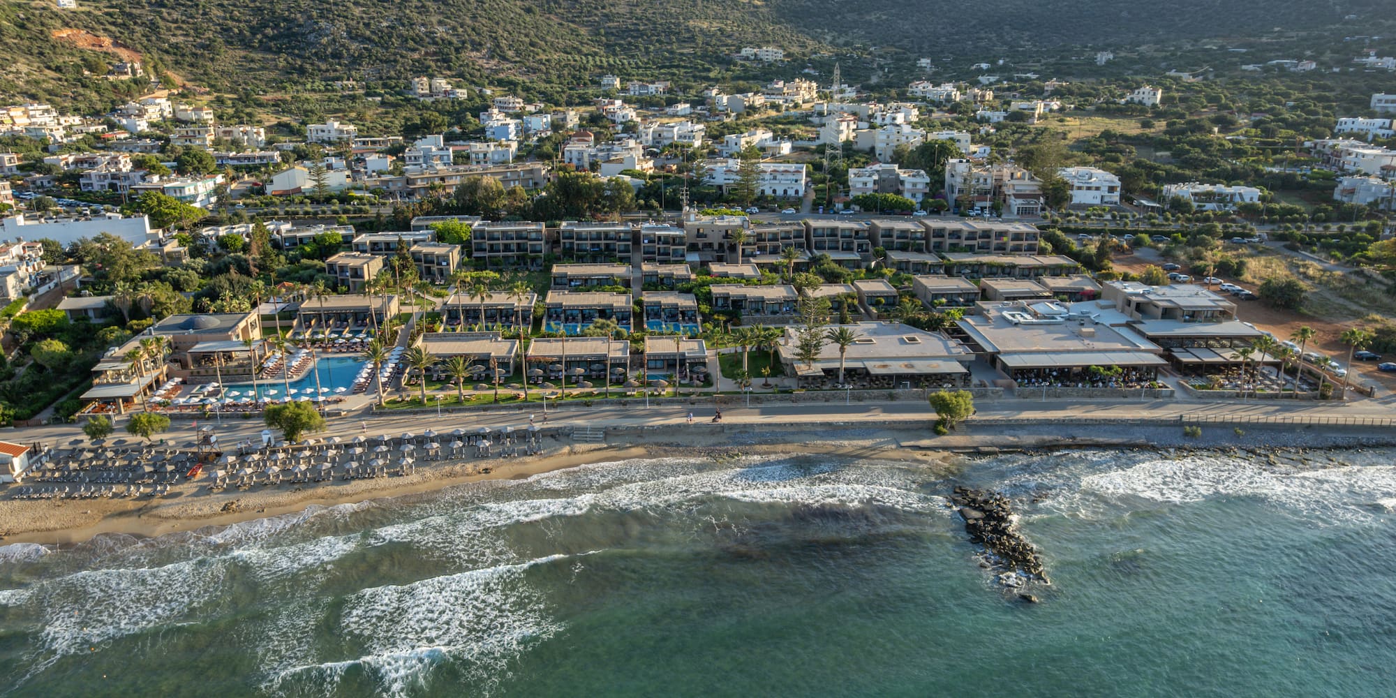 Coastal resort with beach, ocean waves, pools, and green mountains.