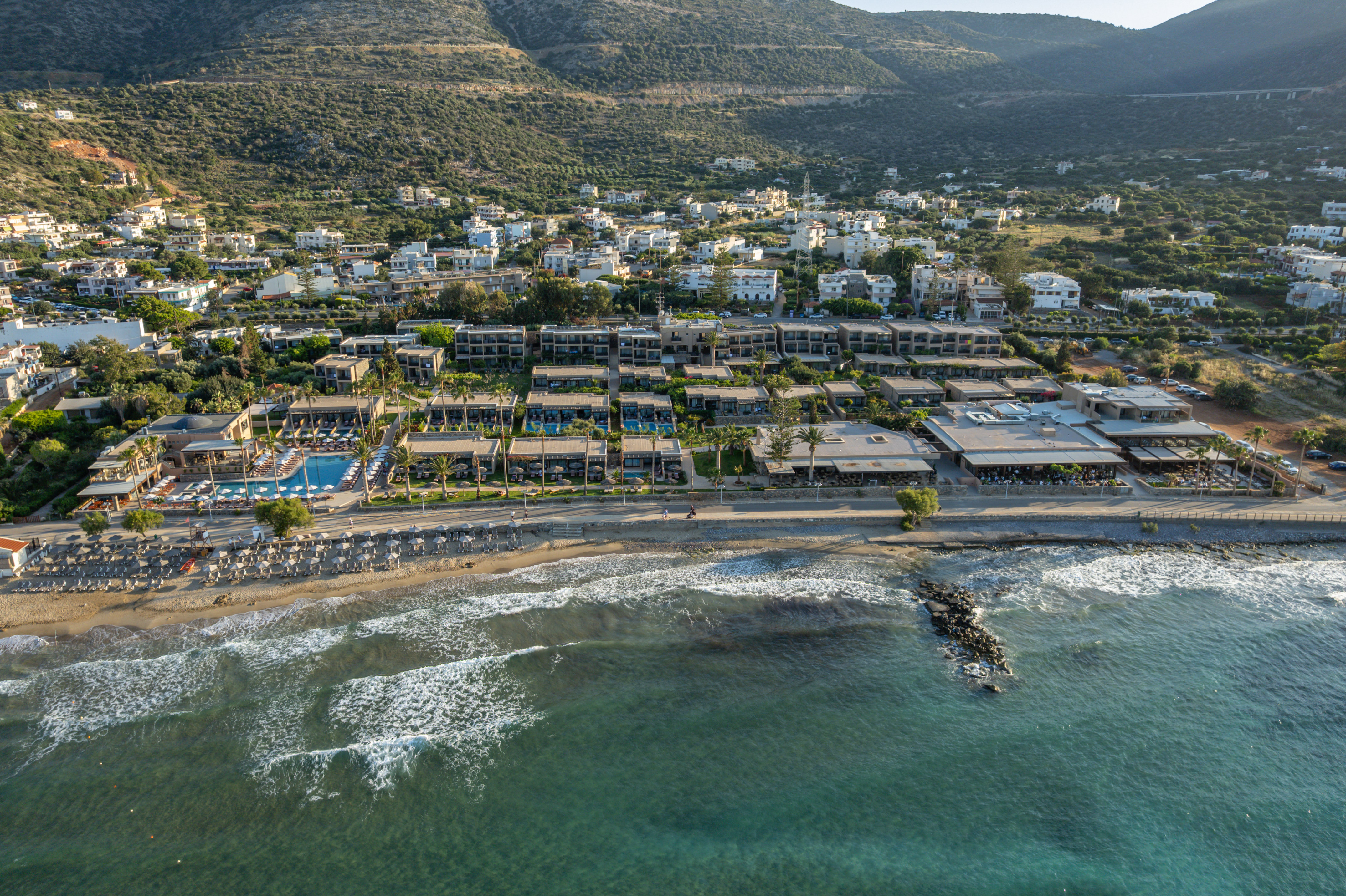 Aerial view: Beachfront resort with pools, sandy beach, clear ocean, and mountains.