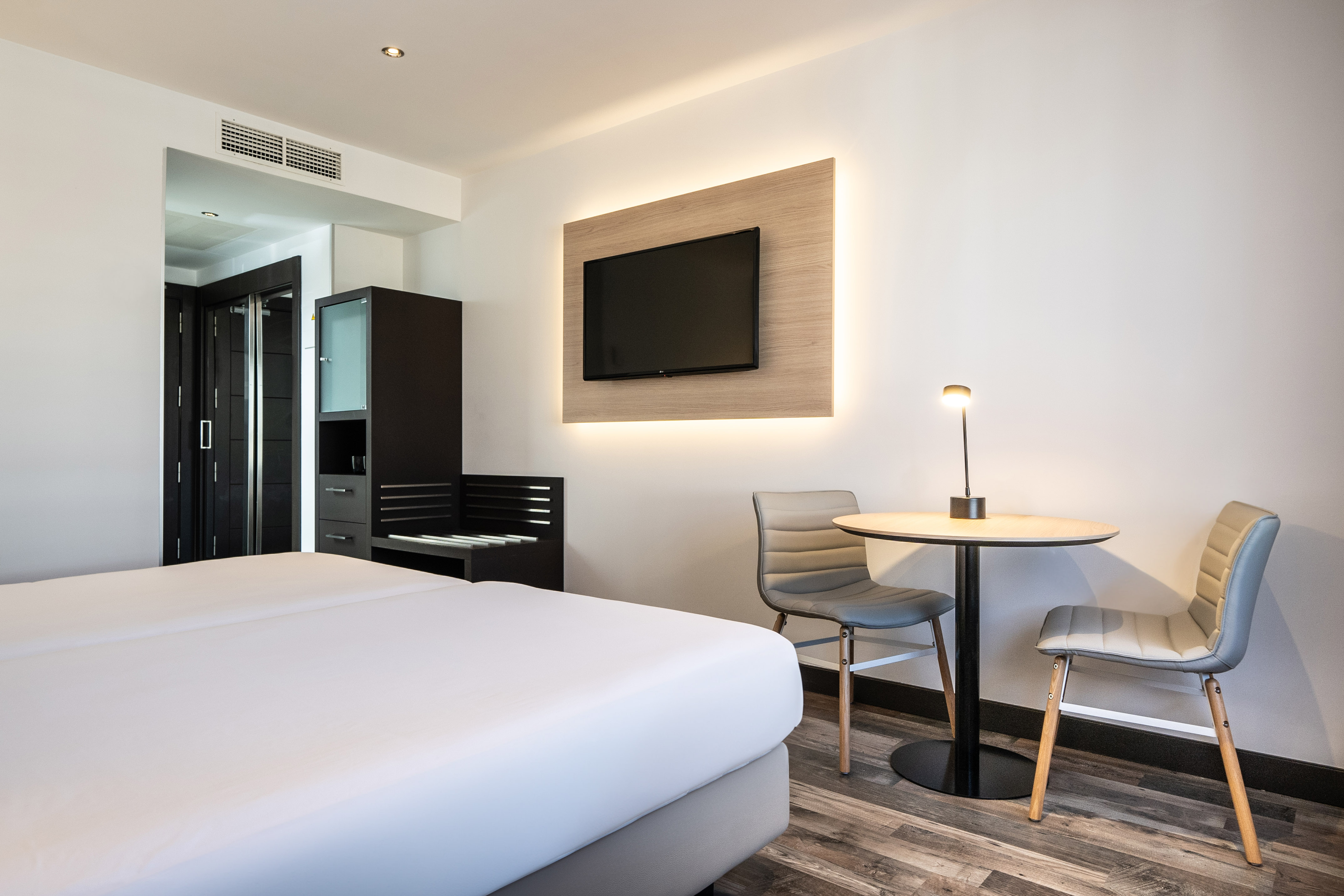 Modern hotel guest room with white bed, backlit TV, and sitting area.