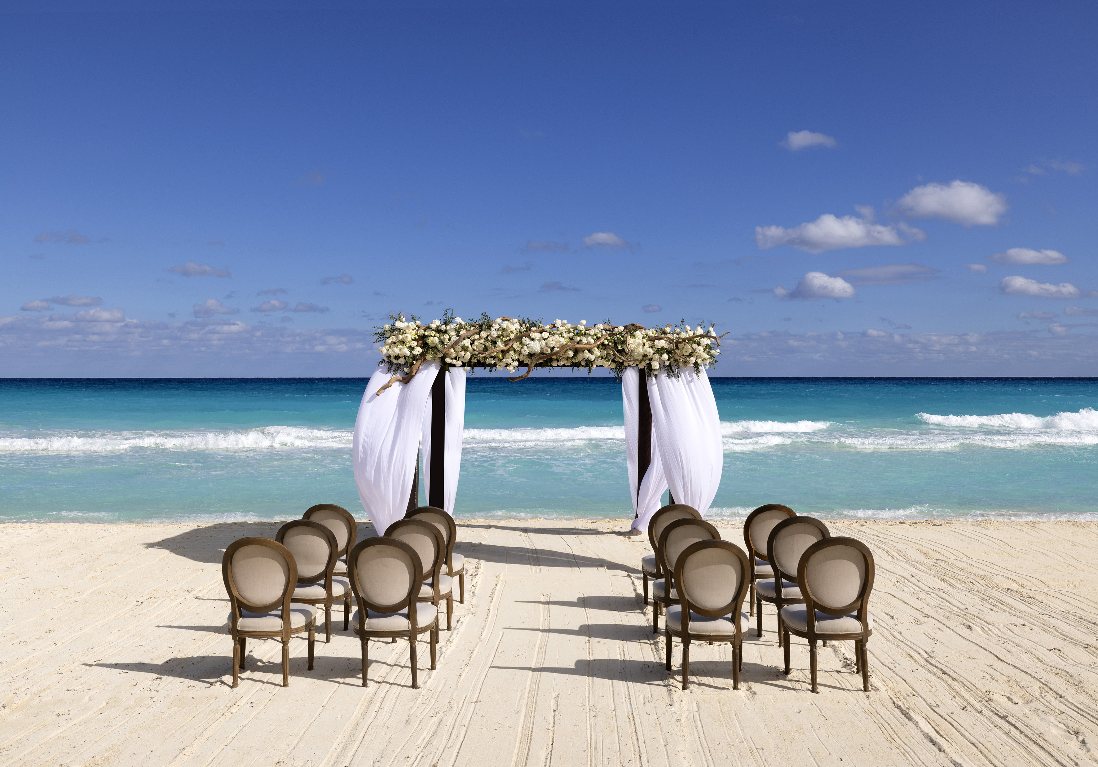 Beach wedding ceremony on sand with floral arch and chairs facing turquoise ocean.
