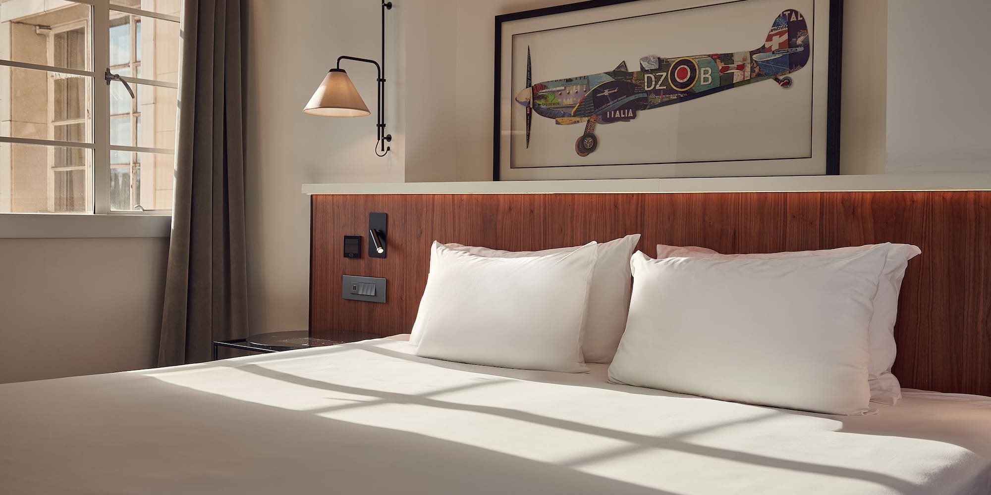 Modern hotel bedroom with white bed, wooden headboard, aviation art, and natural light.
