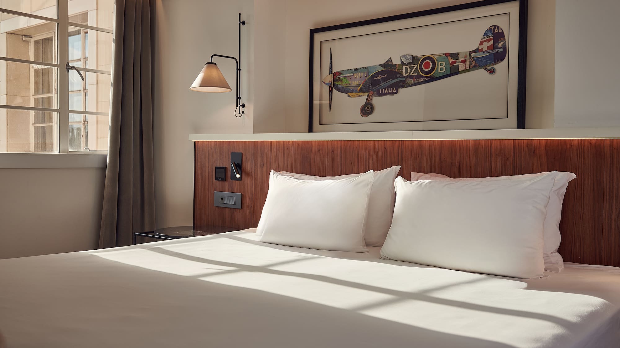 Modern hotel bedroom with white bed, wooden headboard, aviation art, and natural light.