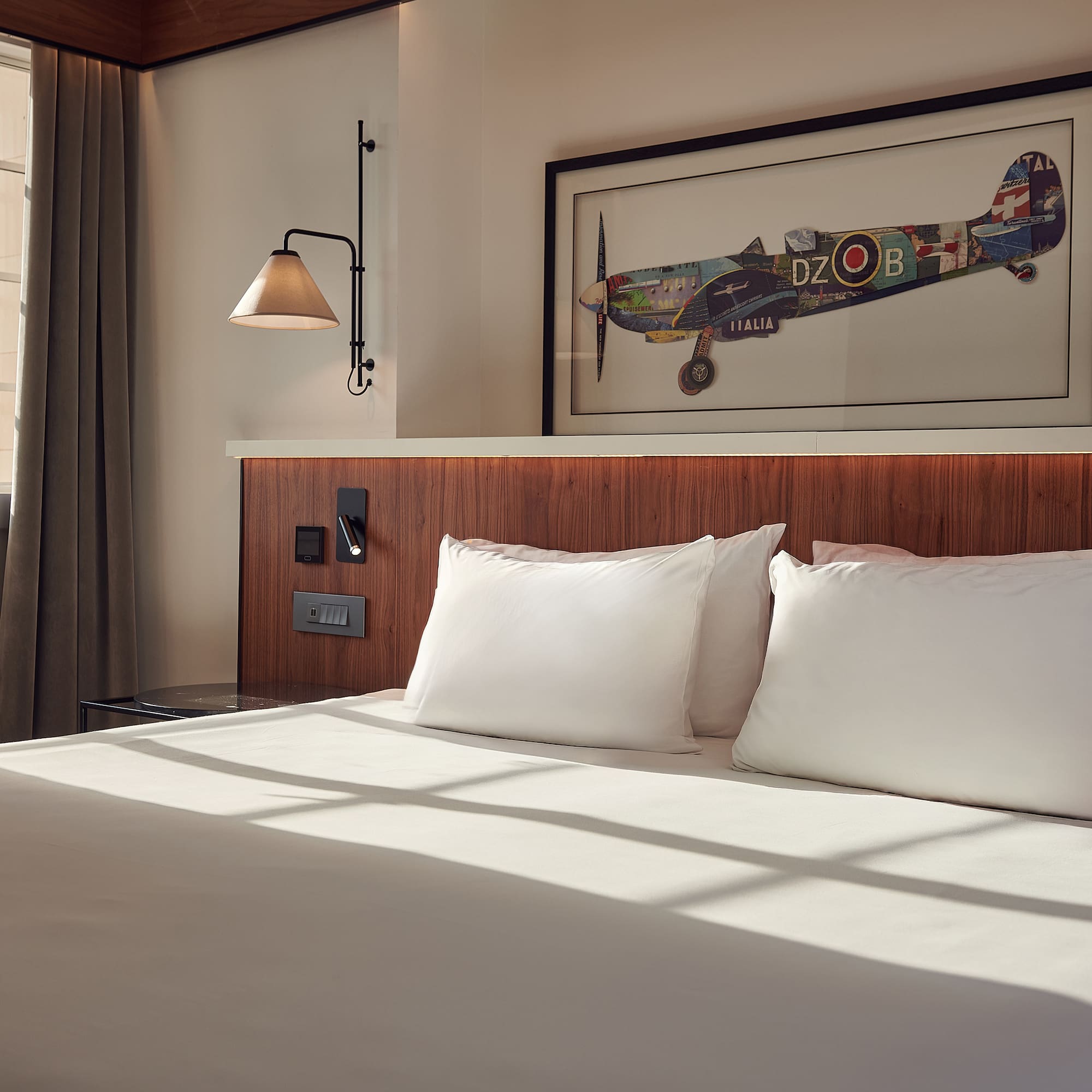 Modern hotel bedroom with white bed, wooden headboard, aviation art, and natural light.