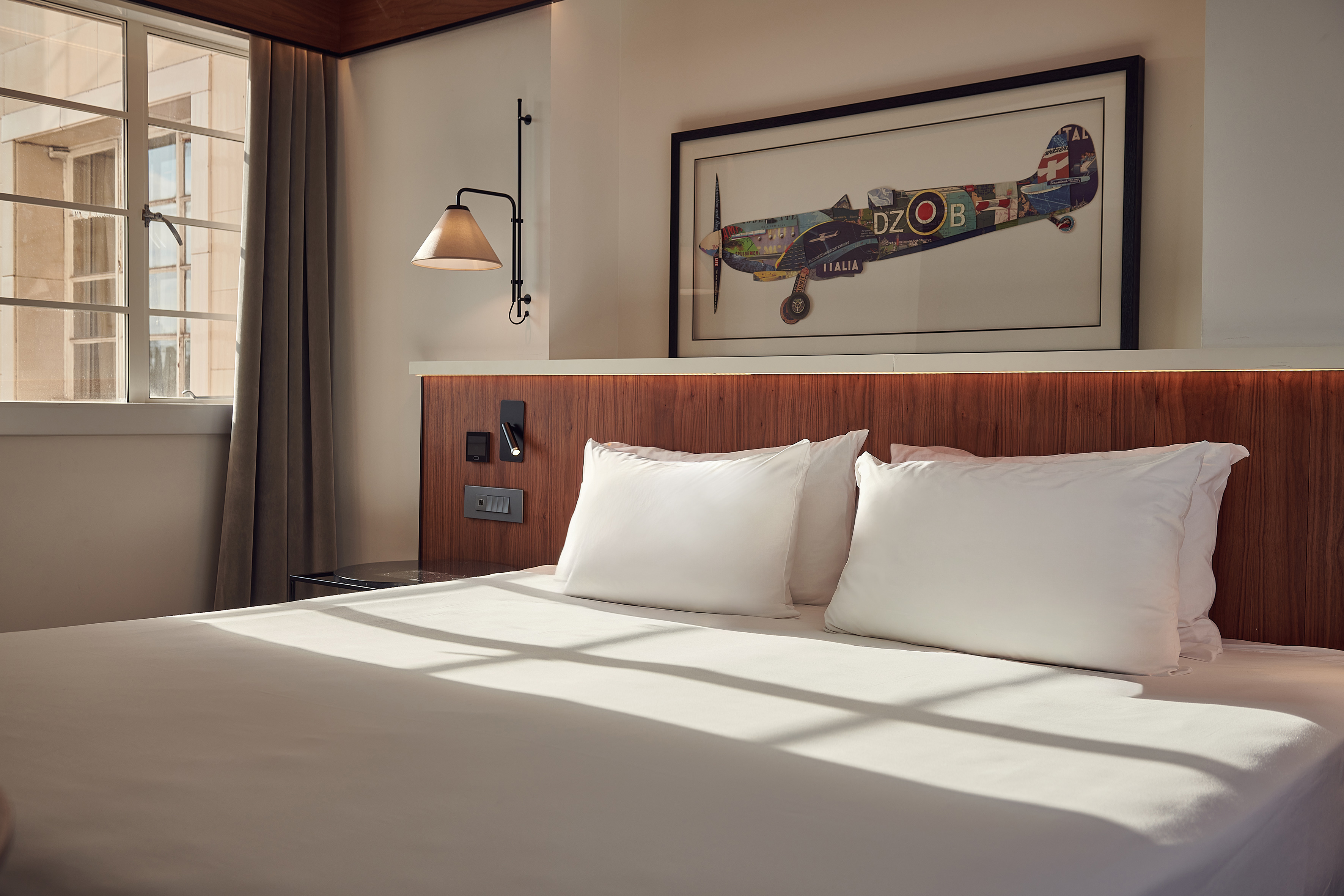 Modern hotel bedroom with white bed, wooden headboard, aviation art, and natural light.