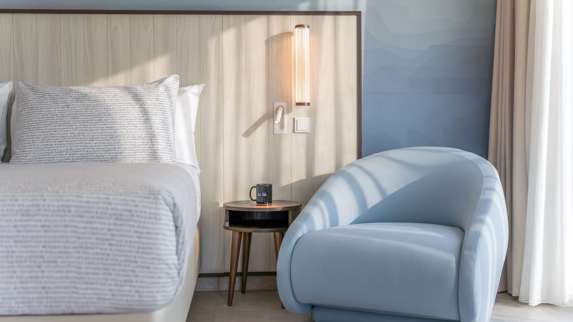 Modern hotel bedroom with light blue armchair, bed, and wooden headboard.