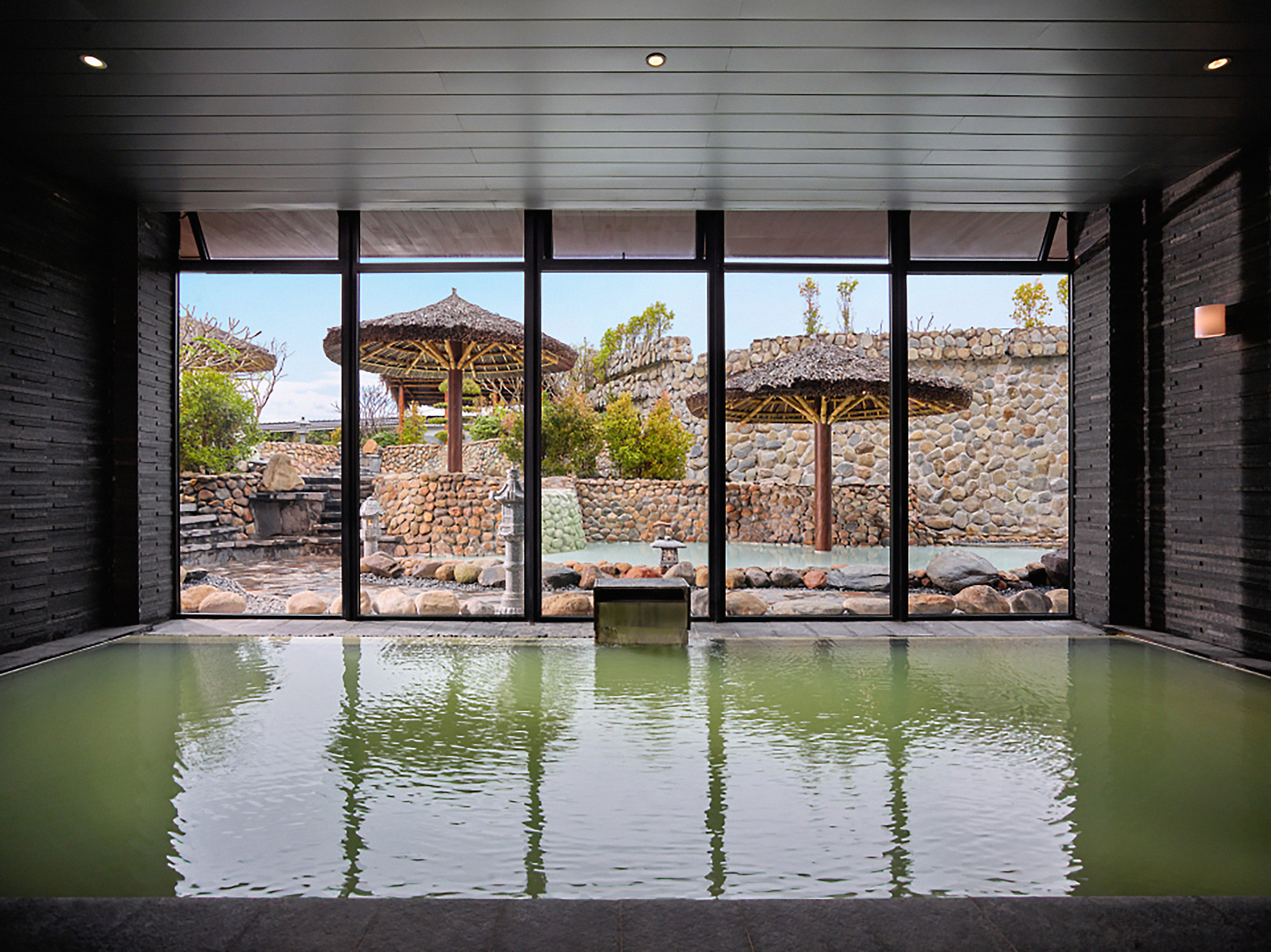 Indoor onsen overlooking an outdoor Japanese garden with thatched gazebos.
