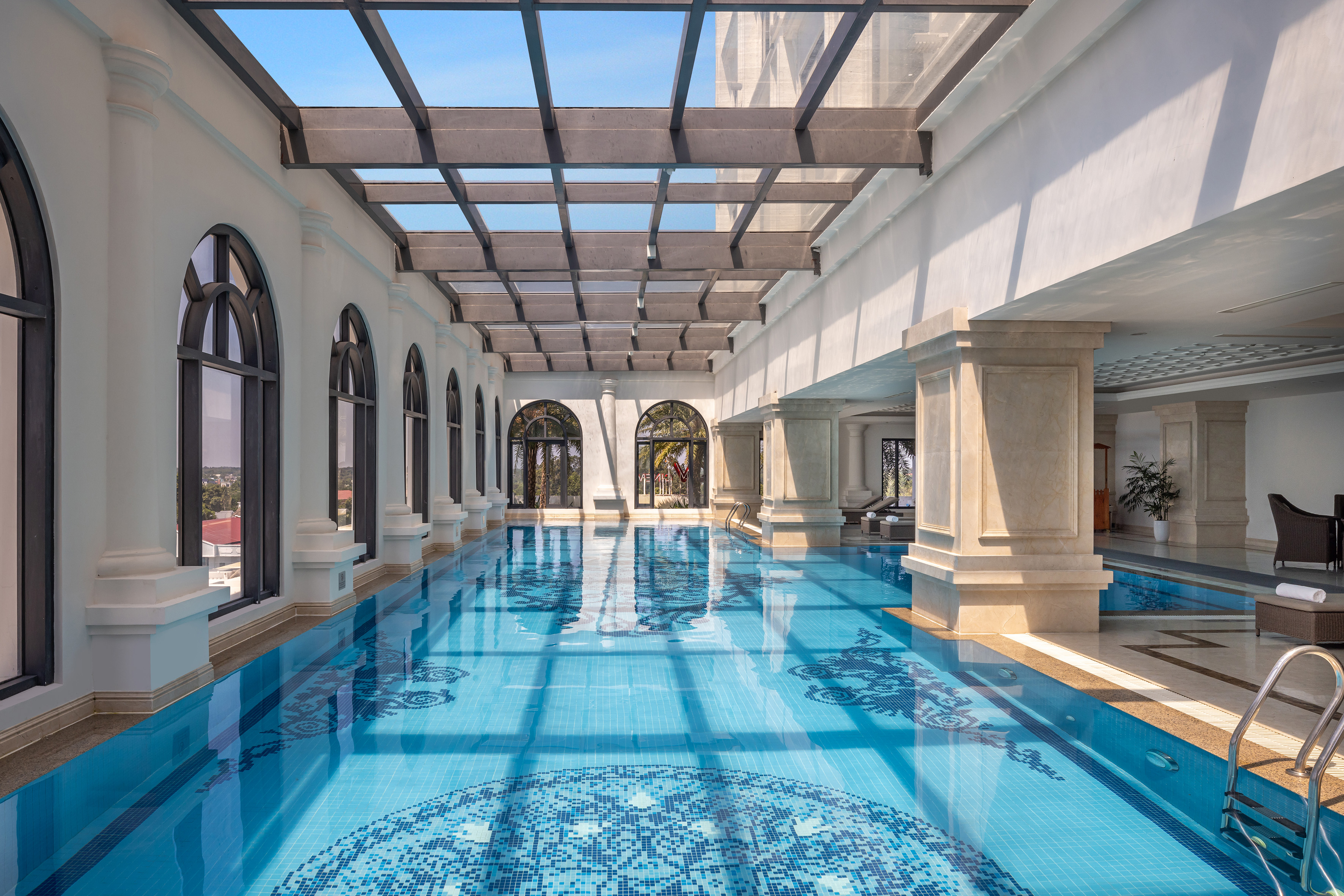 Indoor hotel pool with glass roof, arched windows, and blue mosaic tiles.