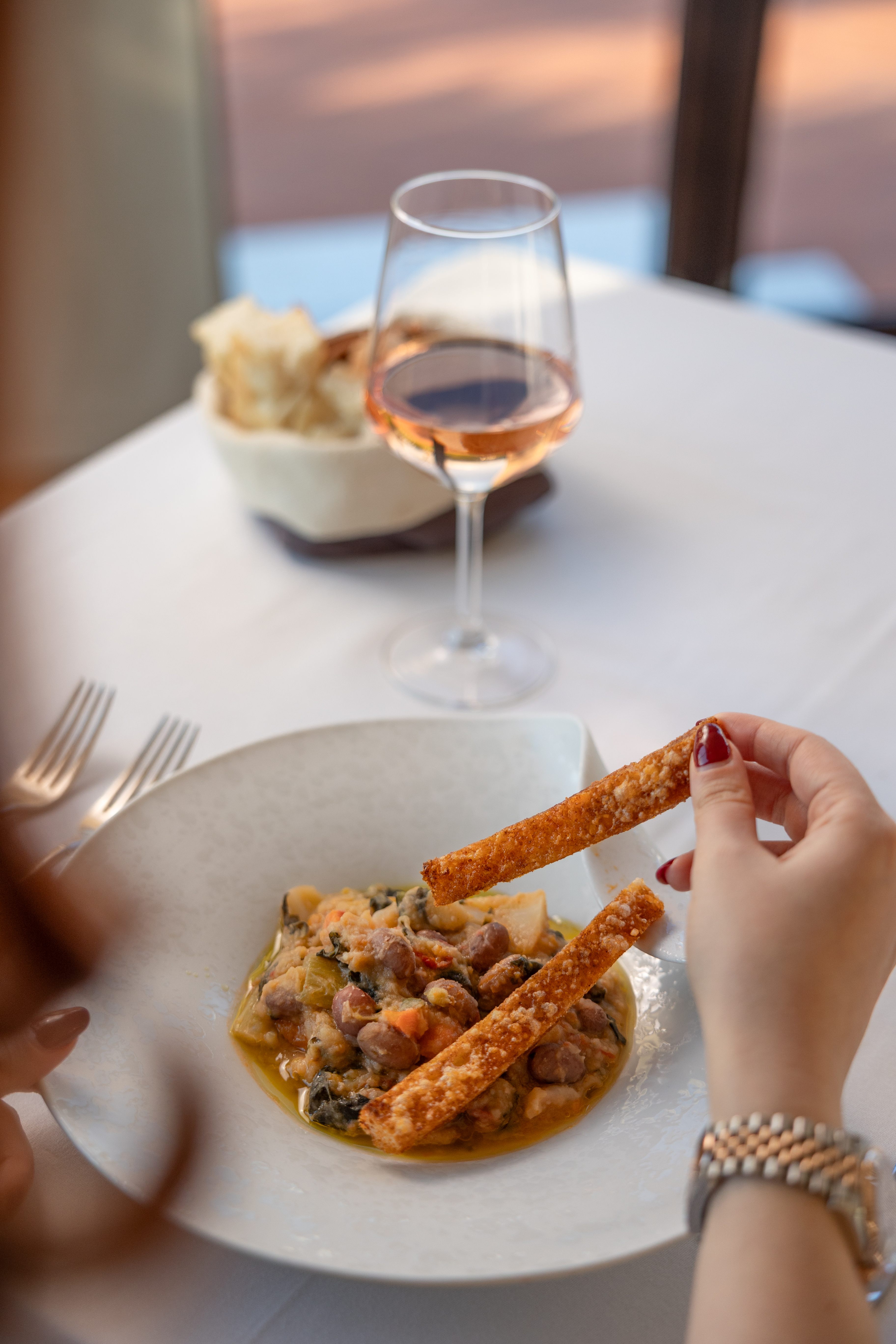 Hand dipping breadstick into bean stew; rosé wine, restaurant meal.