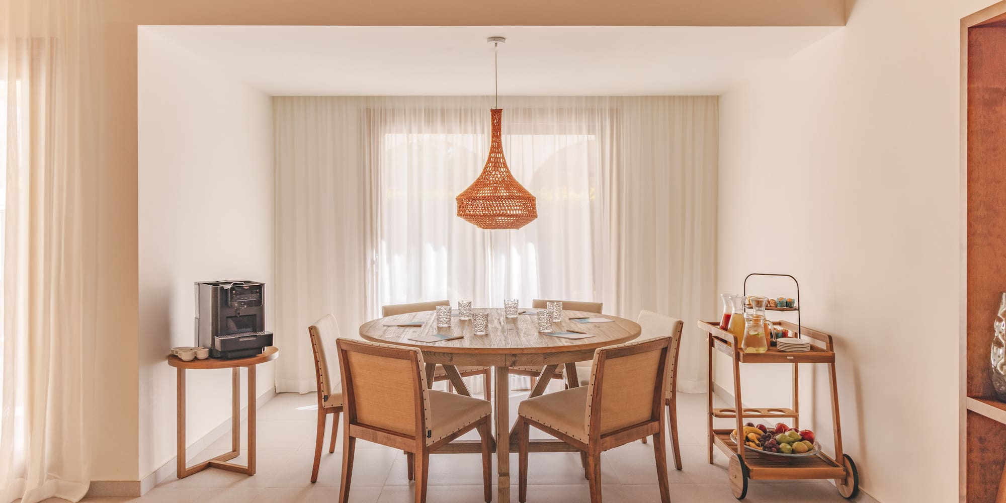 Dining area with wooden table, woven light, coffee, and breakfast cart.