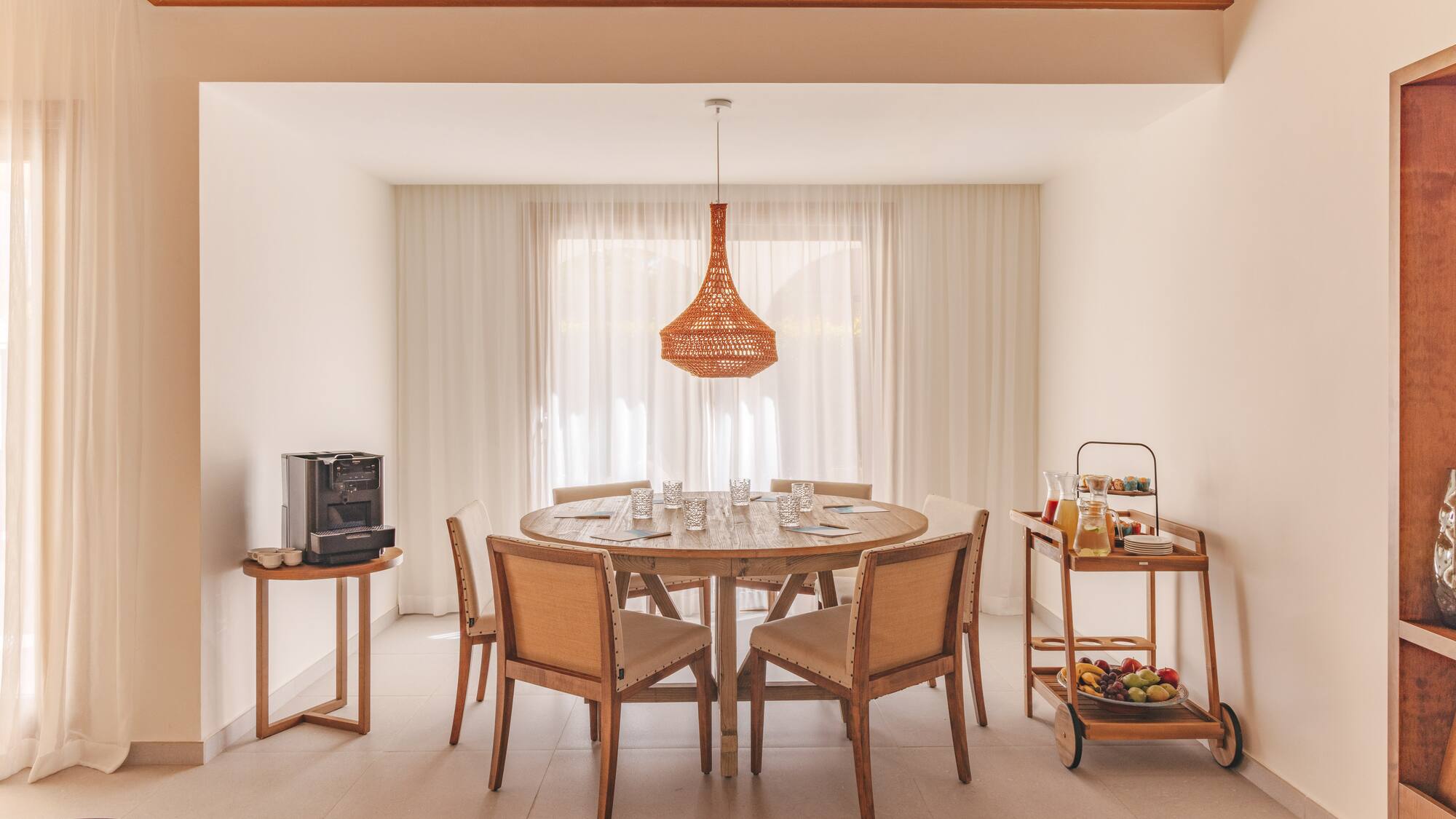 Dining area with wooden table, woven light, coffee, and breakfast cart.