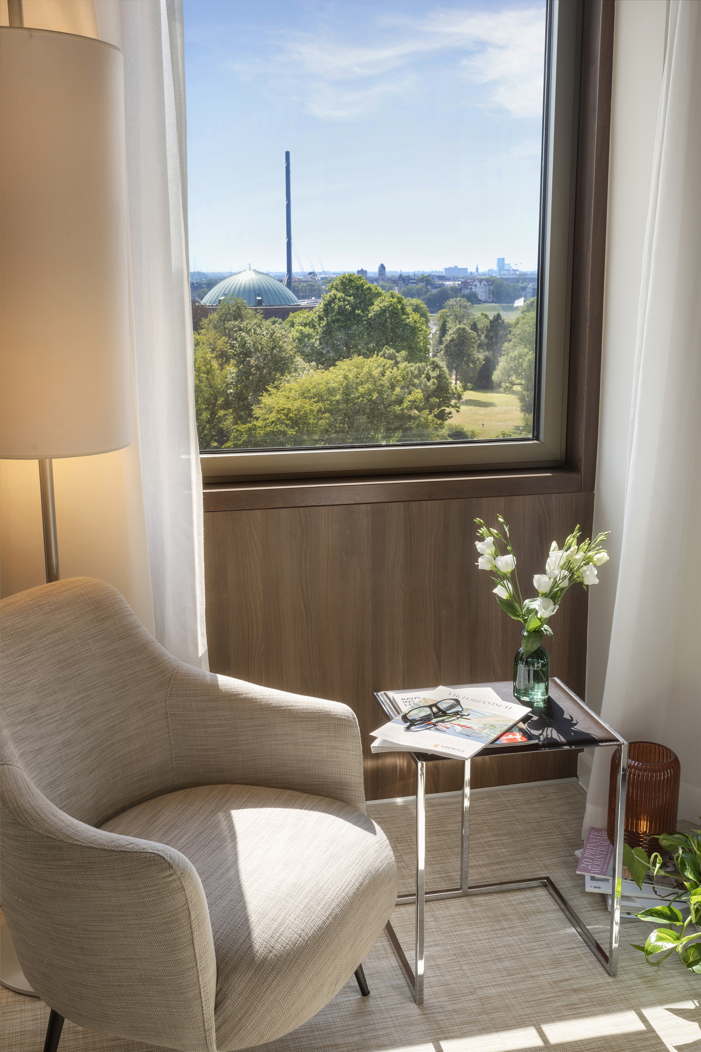 Bright hotel room with comfy armchair, flowers, and Düsseldorf city park view with Tonhalle.