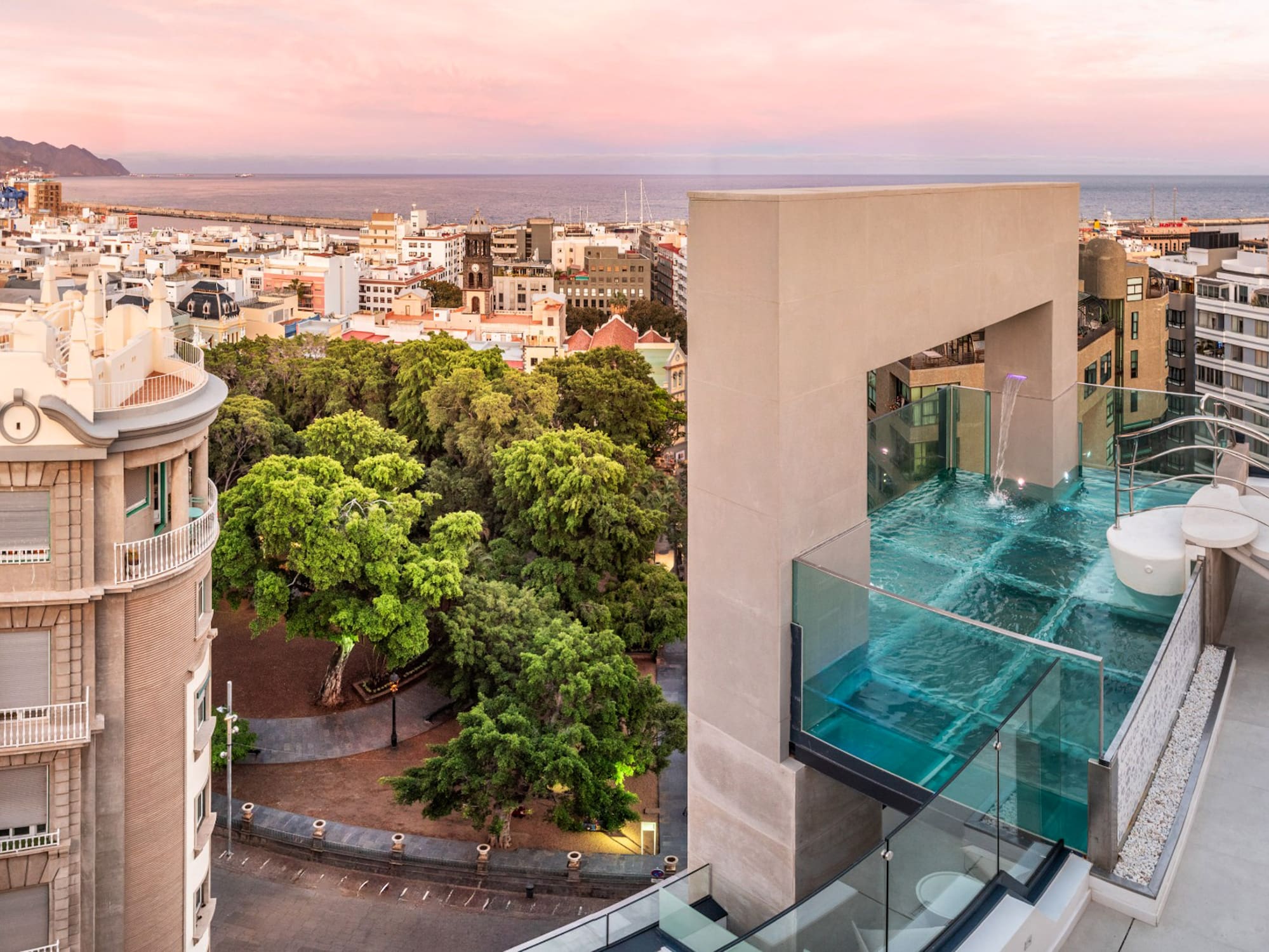 Glass-walled rooftop infinity pool with city and ocean view at pink sunset.
