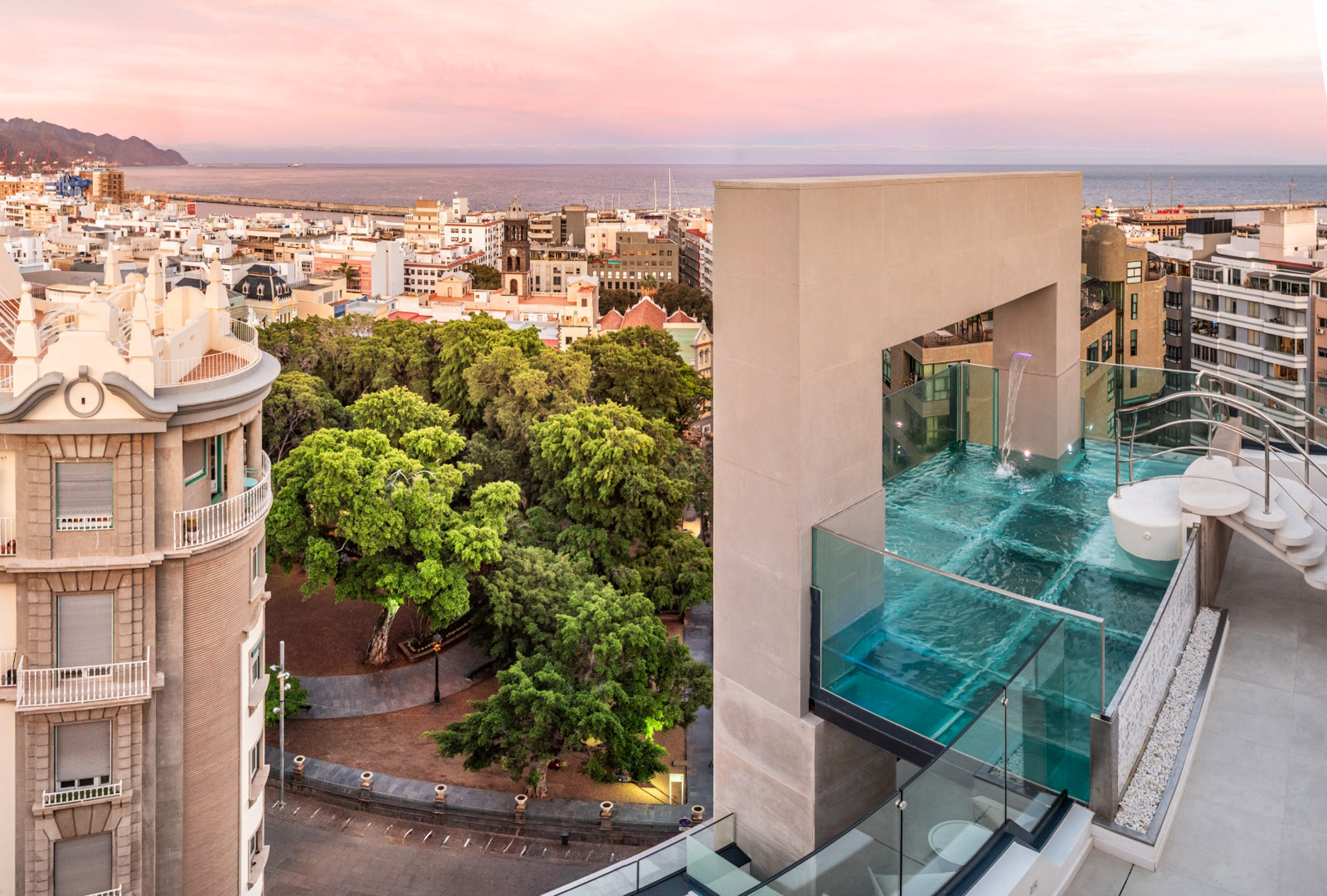 Glass-walled rooftop infinity pool with city and ocean view at pink sunset.