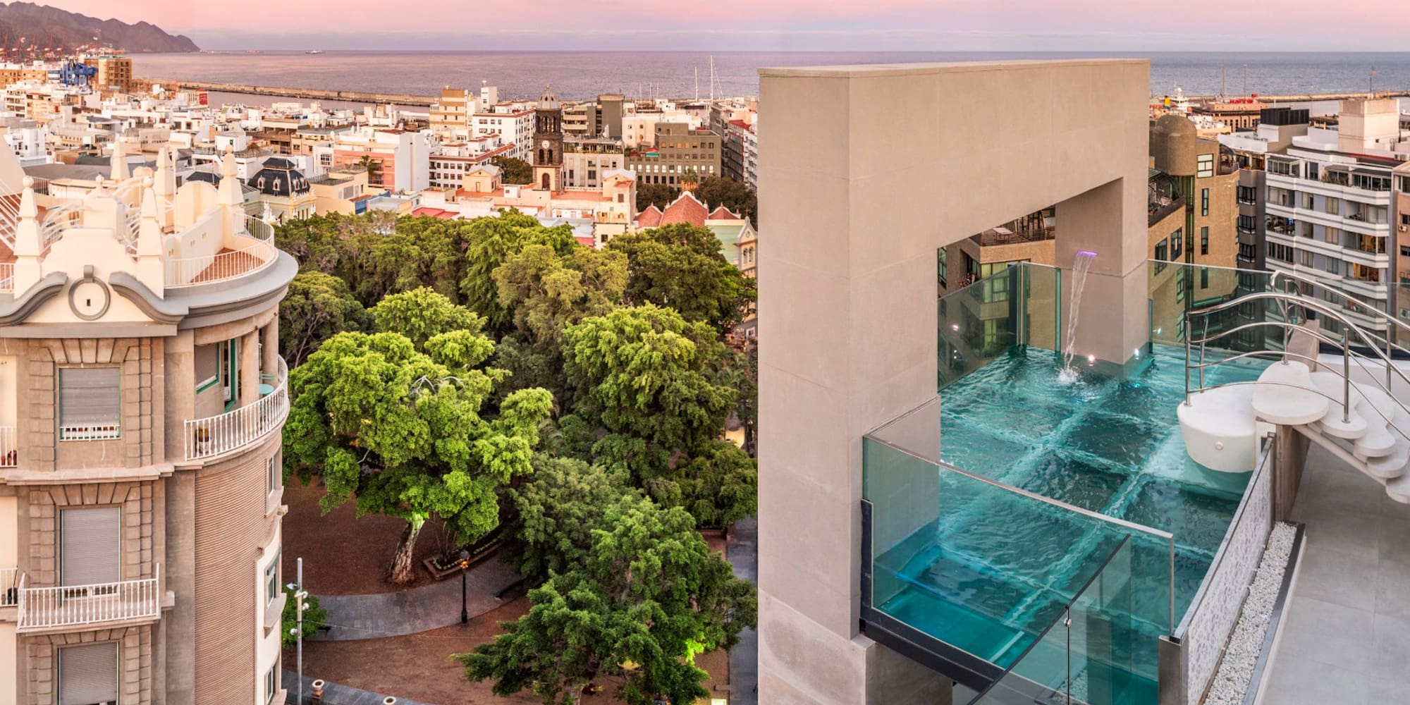 Glass-walled rooftop infinity pool with city and ocean view at pink sunset.