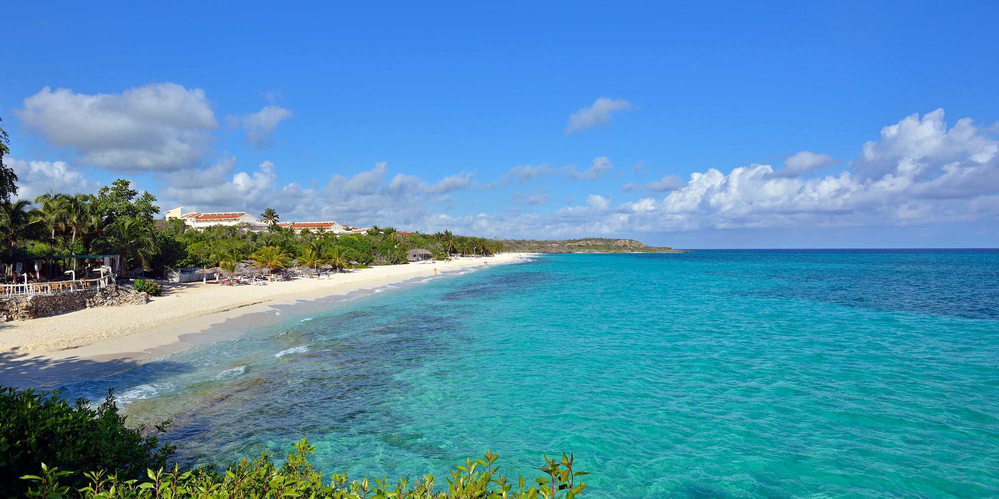 Tropical white sand beach, clear turquoise ocean, palm trees, and beachfront resort.