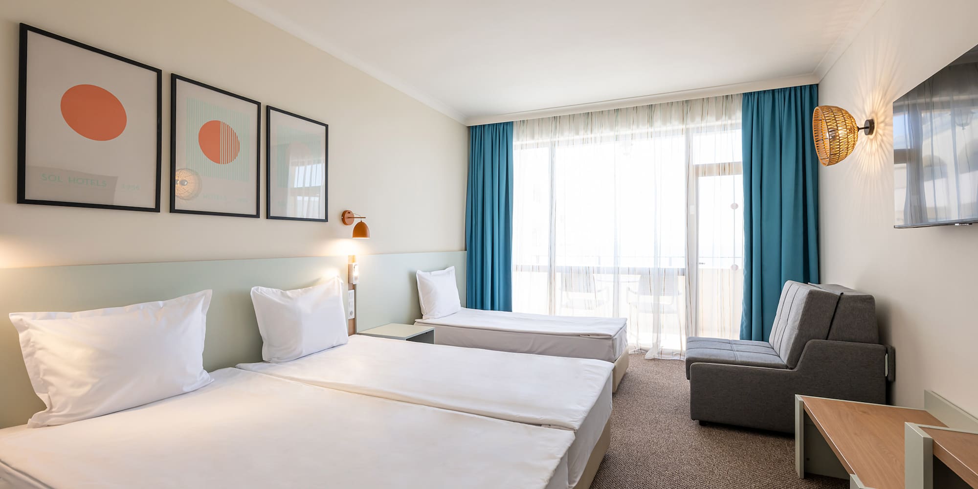 Bright hotel room with three single beds, gray sofa, balcony, and modern art.