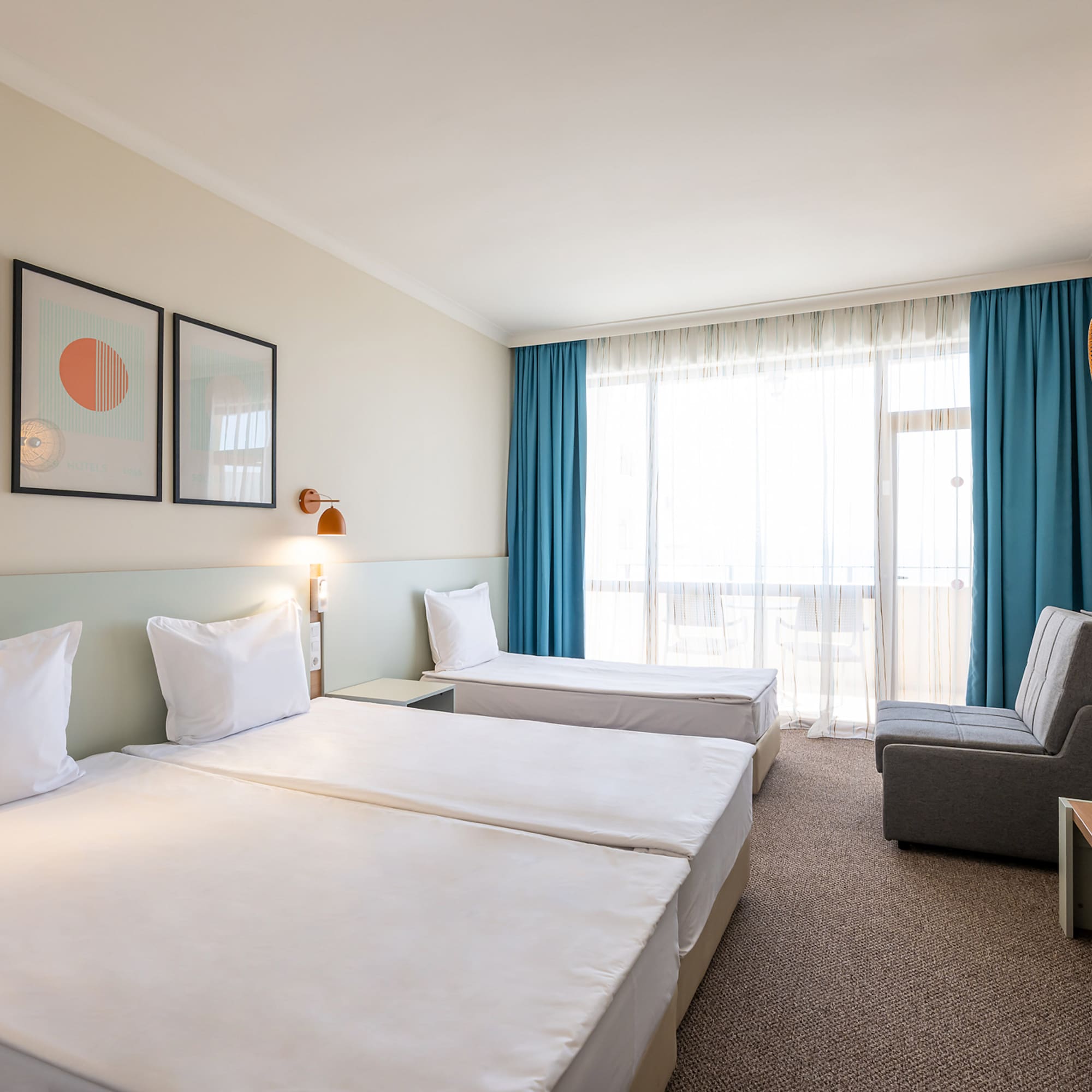 Bright hotel room with three single beds, gray sofa, balcony, and modern art.