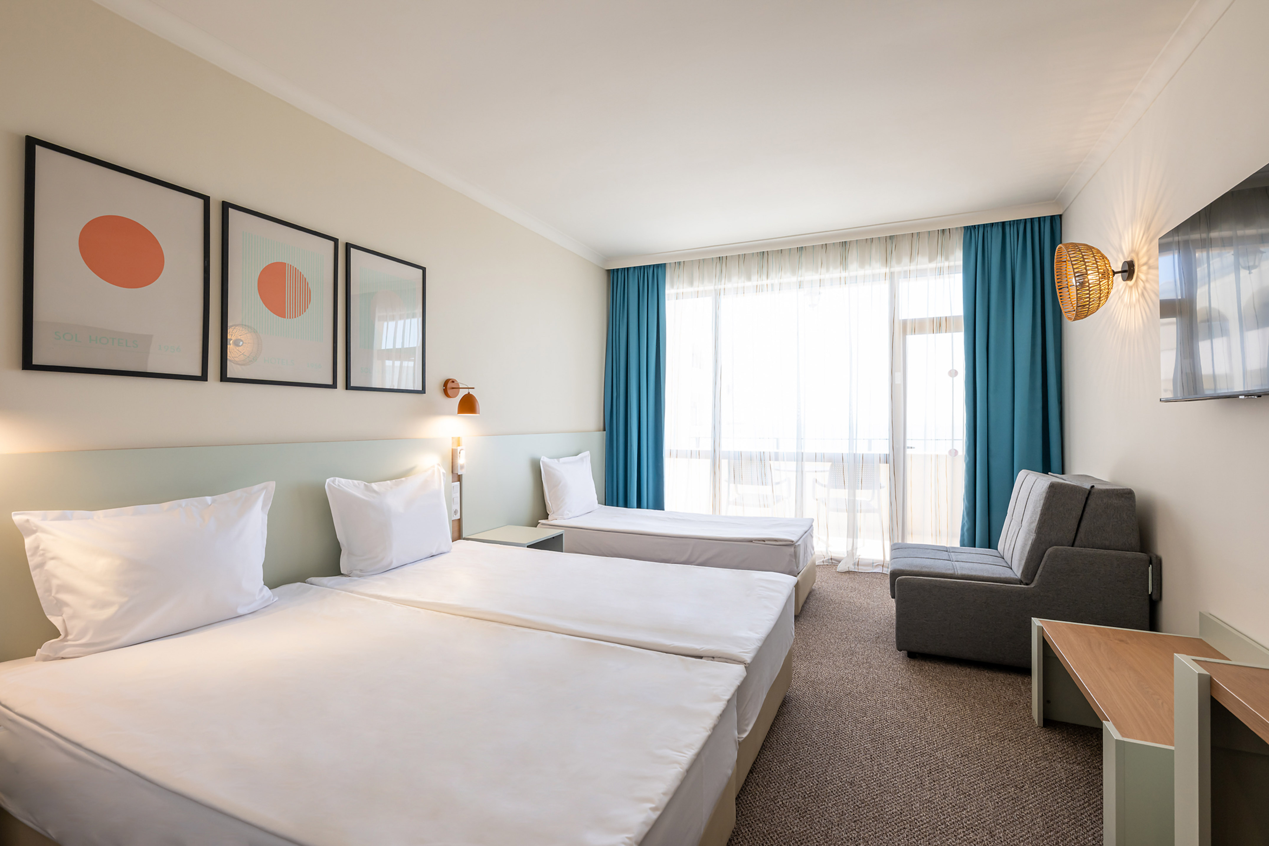 Bright hotel room with three single beds, gray sofa, balcony, and modern art.