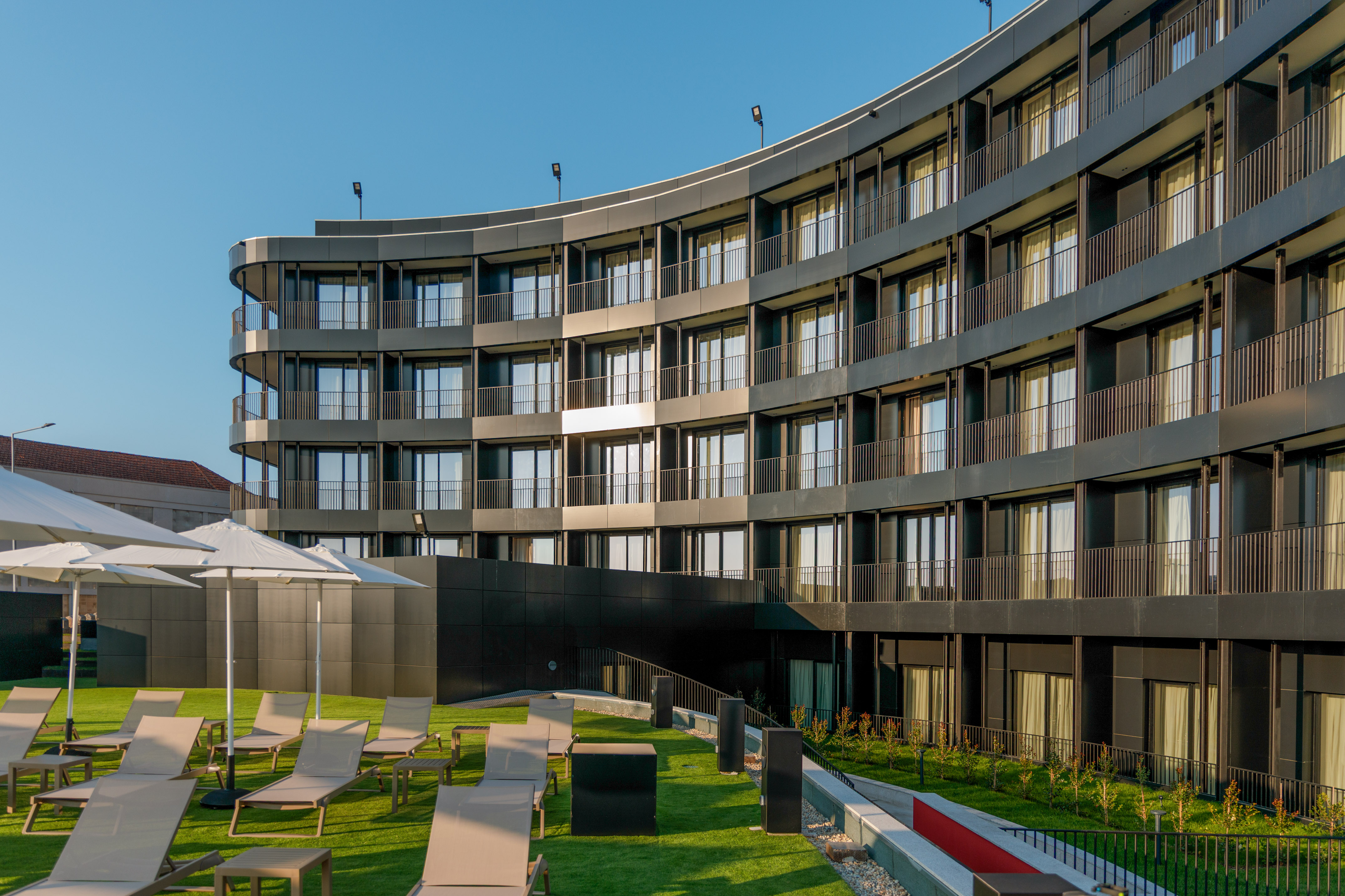 Curved modern hotel building, green terrace with sun loungers & umbrellas.
