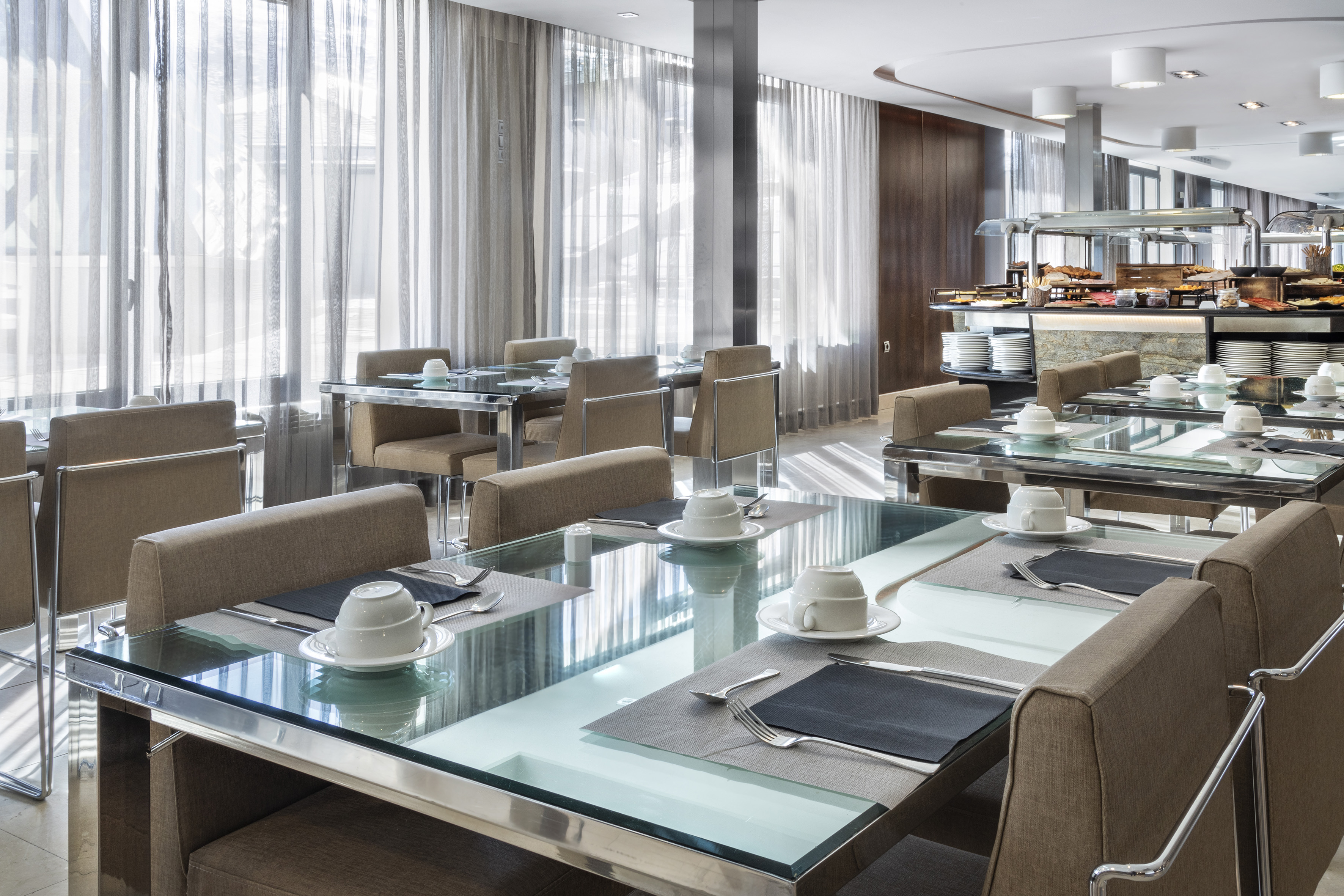 Bright restaurant dining room with glass tables, beige chairs, and breakfast buffet.