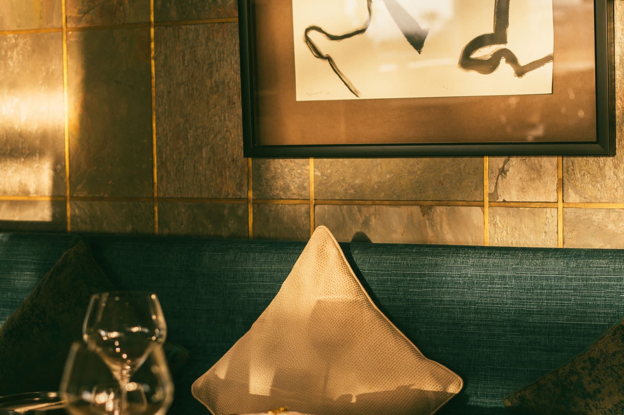 Restaurant table with wine glasses, teal banquette, abstract art on tiled wall.