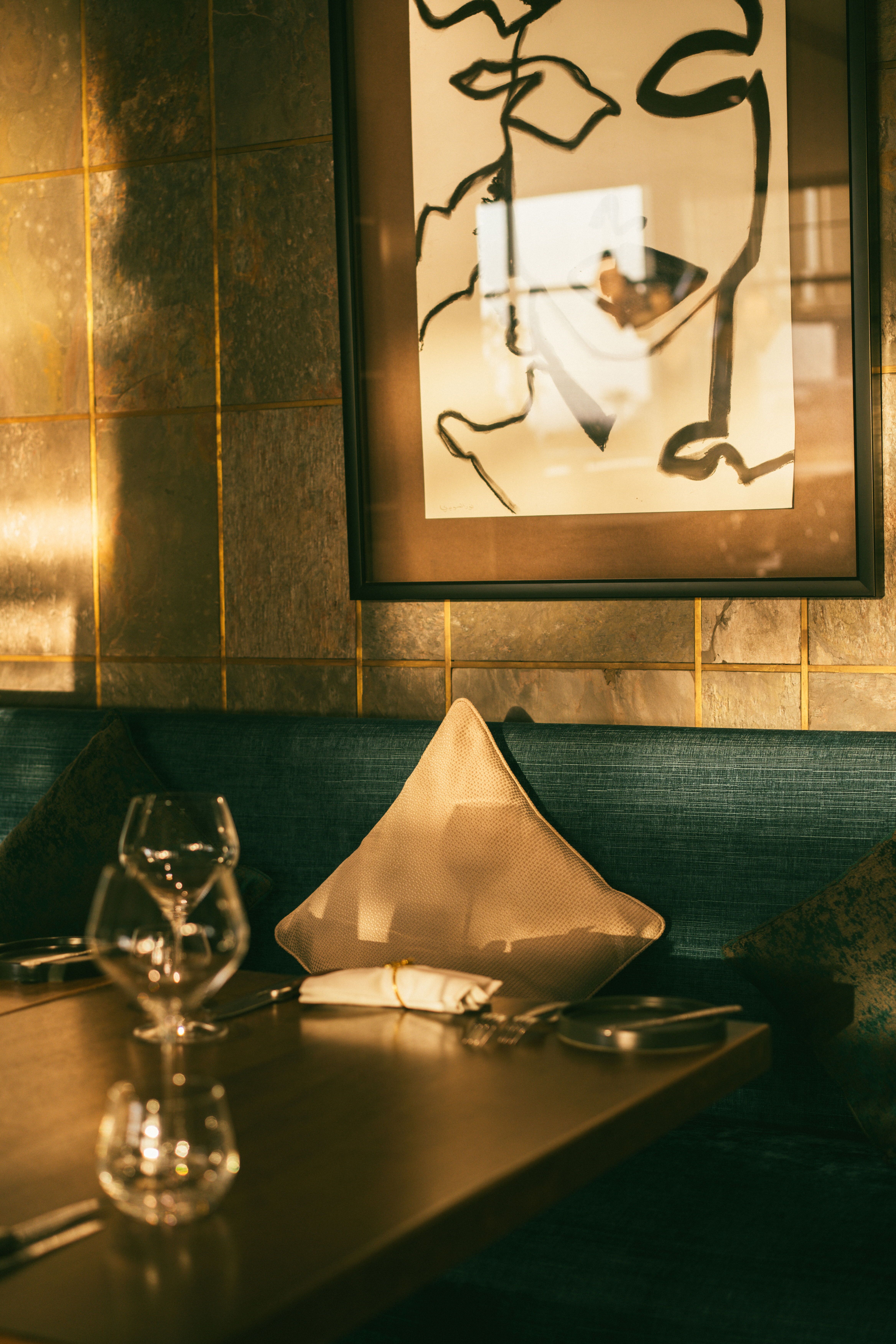 Restaurant table with wine glasses, teal banquette, abstract art on tiled wall.
