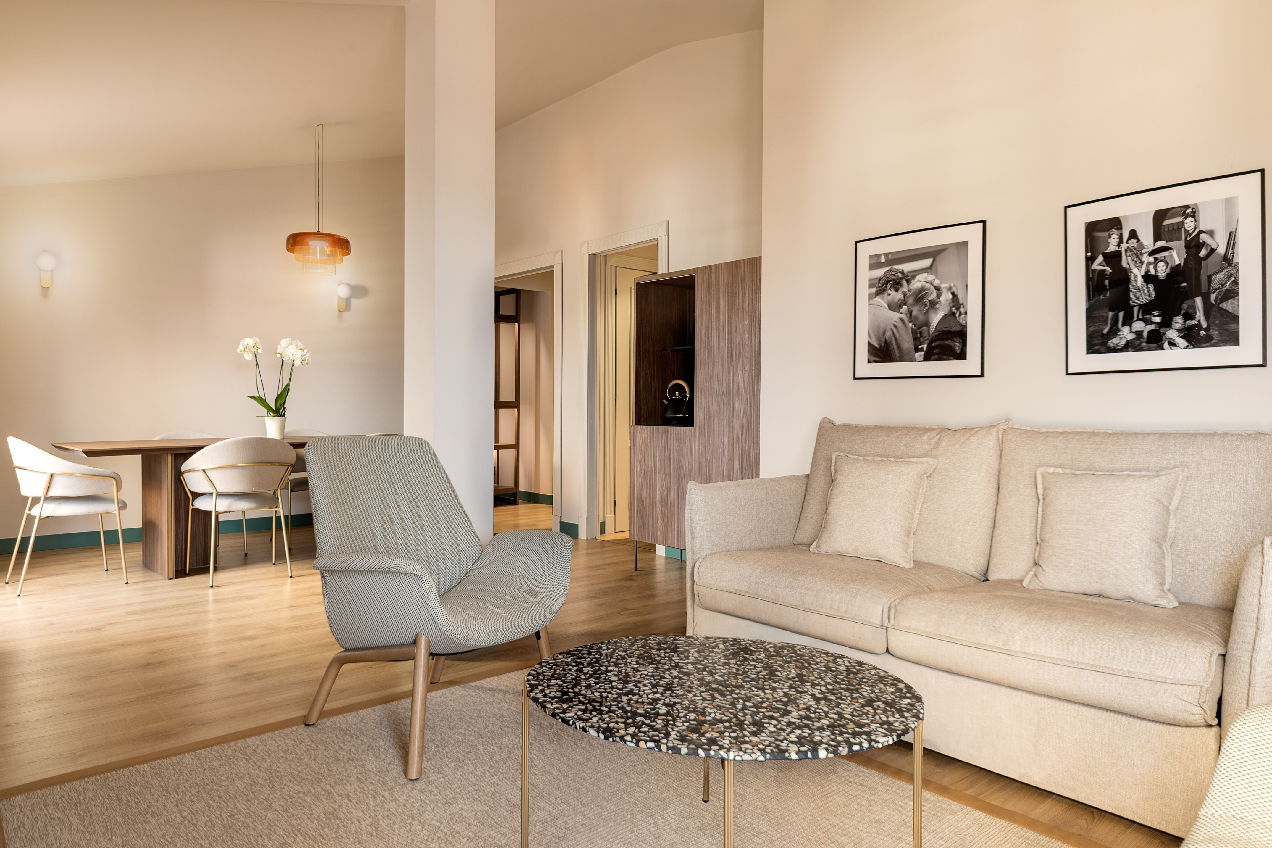 Modern hotel suite living room with sofa, armchair, dining area, and wall art.