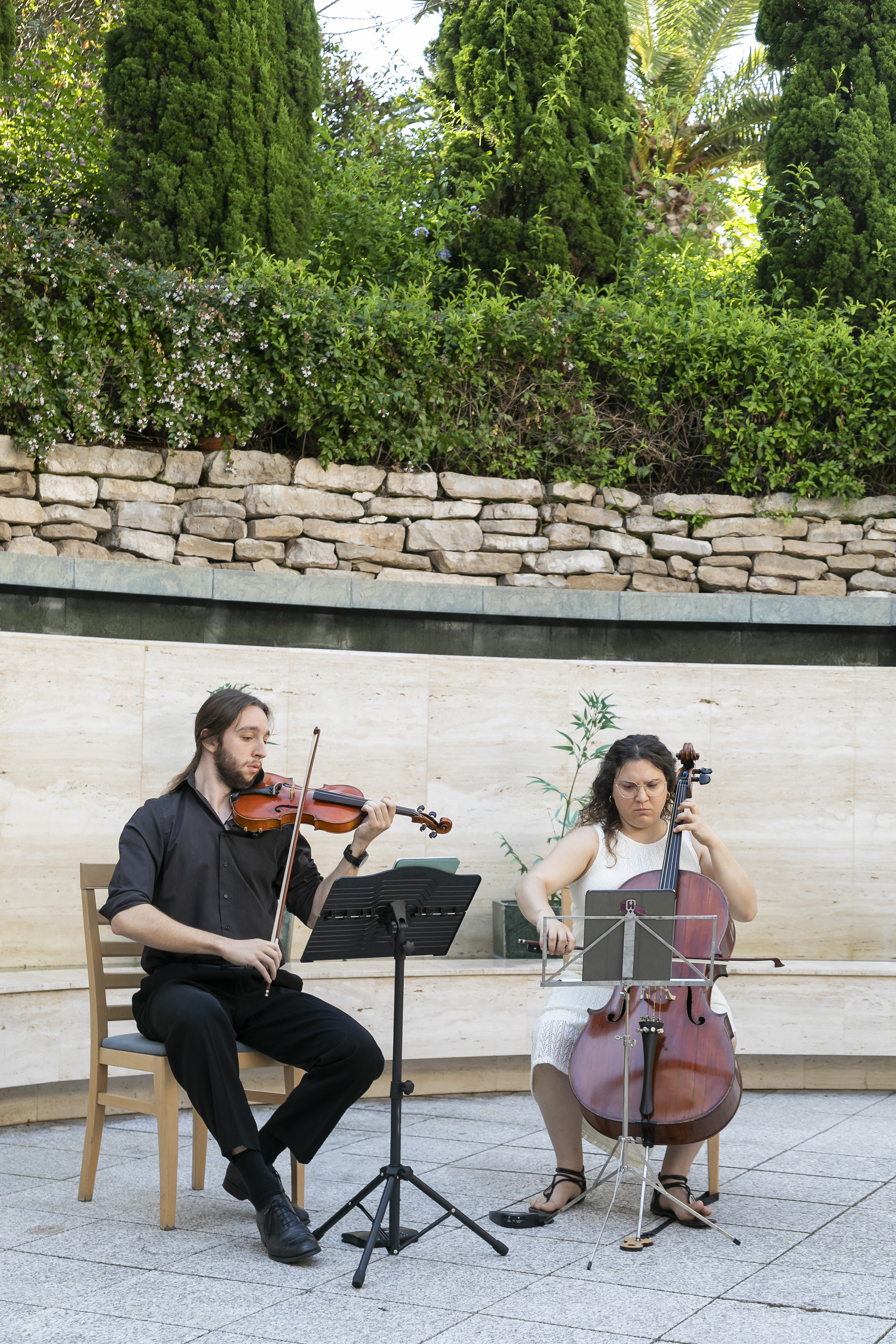 Violinist and cellist performing string instruments outdoors among green plants.