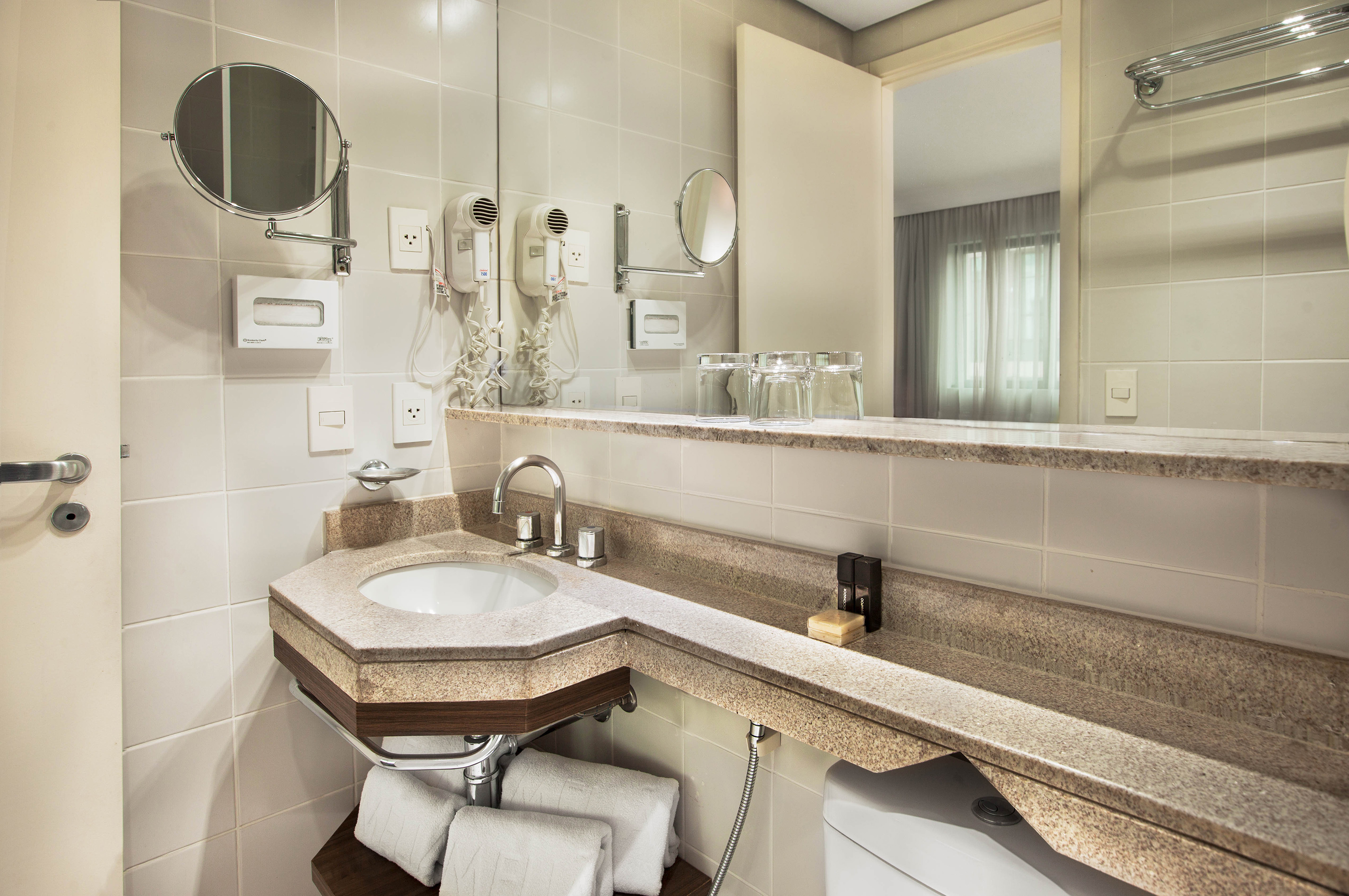 Clean hotel bathroom with stone vanity, sink, mirror, hair dryers, and fresh towels.