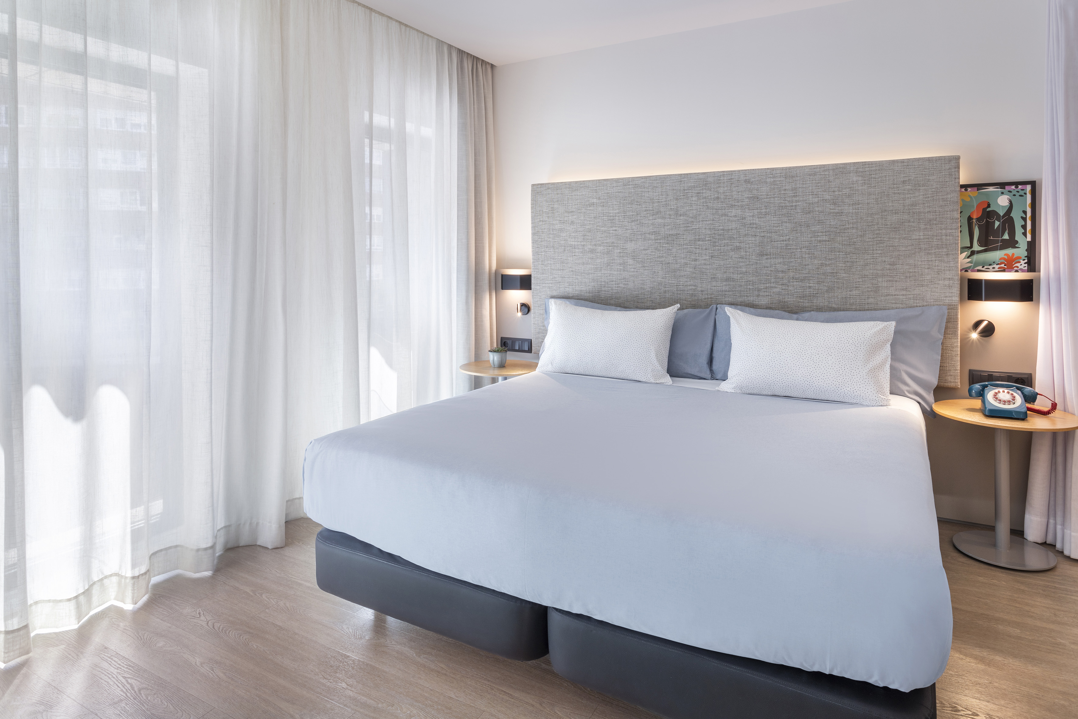 Bright hotel bedroom with grey textured headboard, large bed, and sheer curtains.