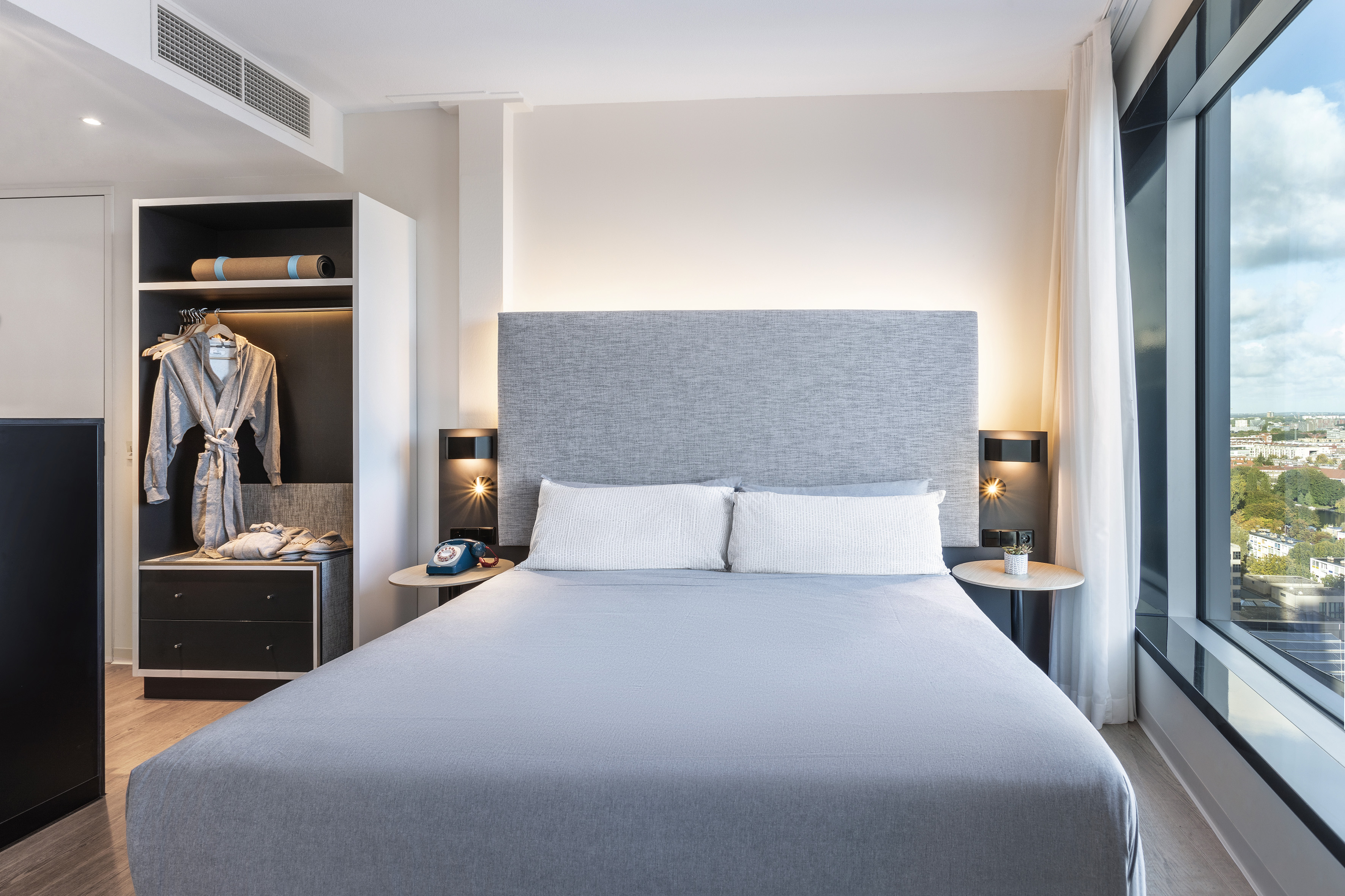 Modern hotel bedroom with grey bed, city view, and yoga mat in closet.