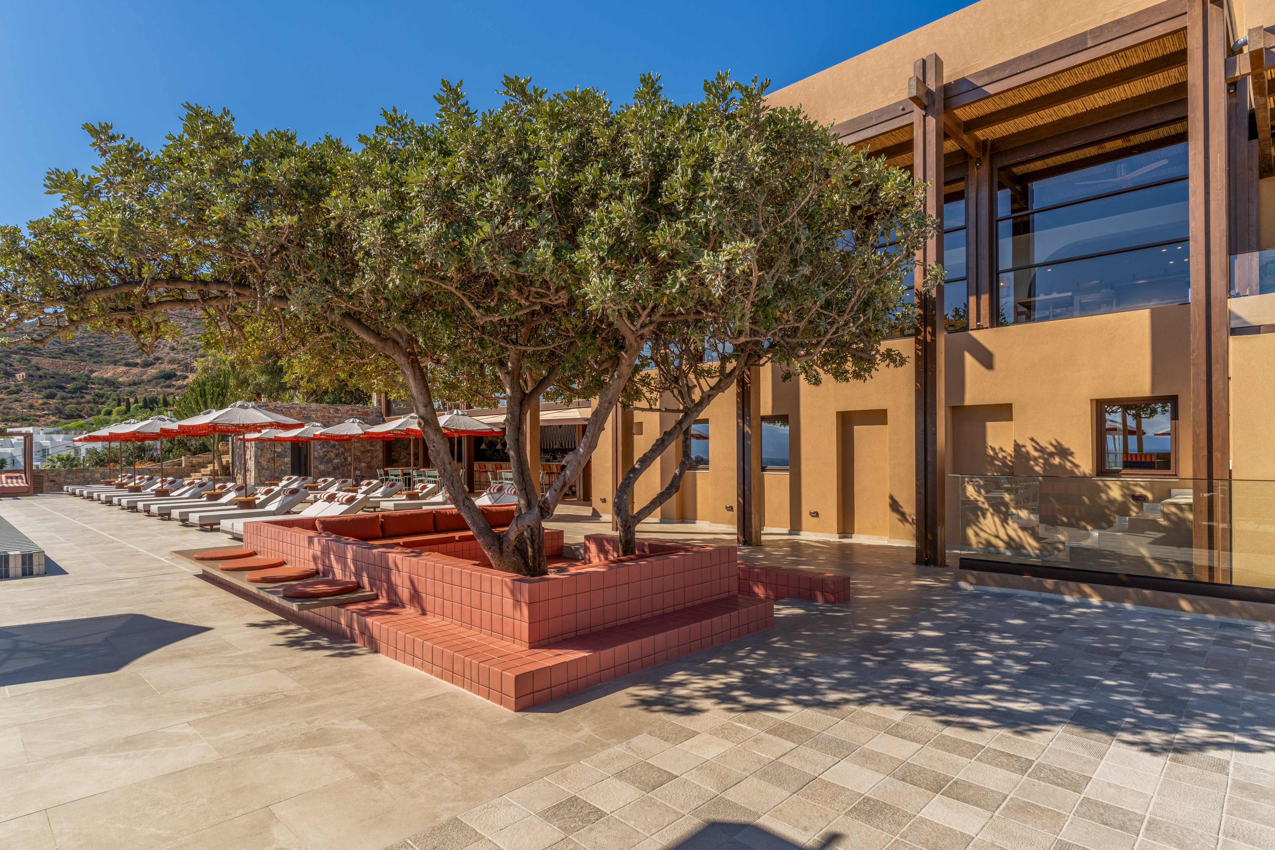 Modern hotel patio with leafy trees, red planters, and sun loungers.