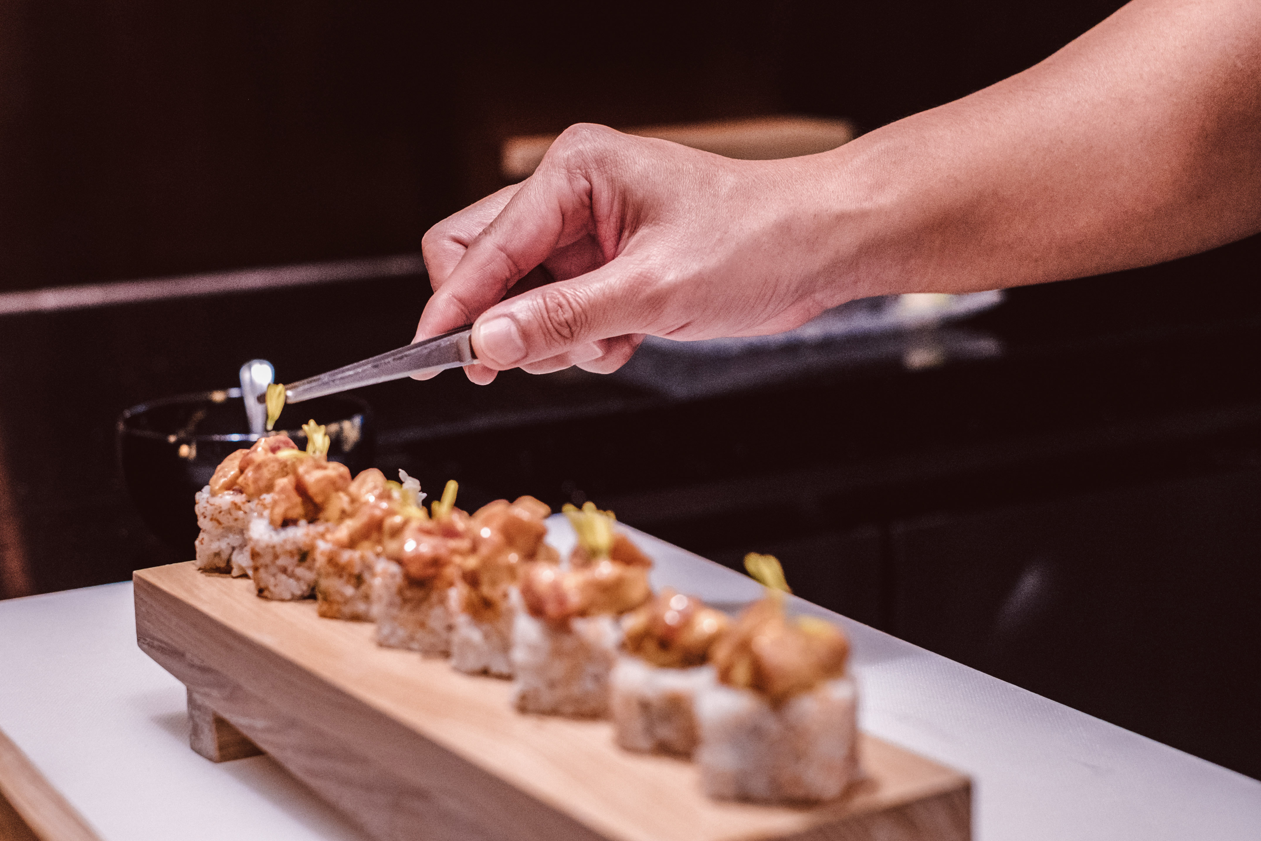 Chef's hand garnishing maki sushi rolls on wooden board.