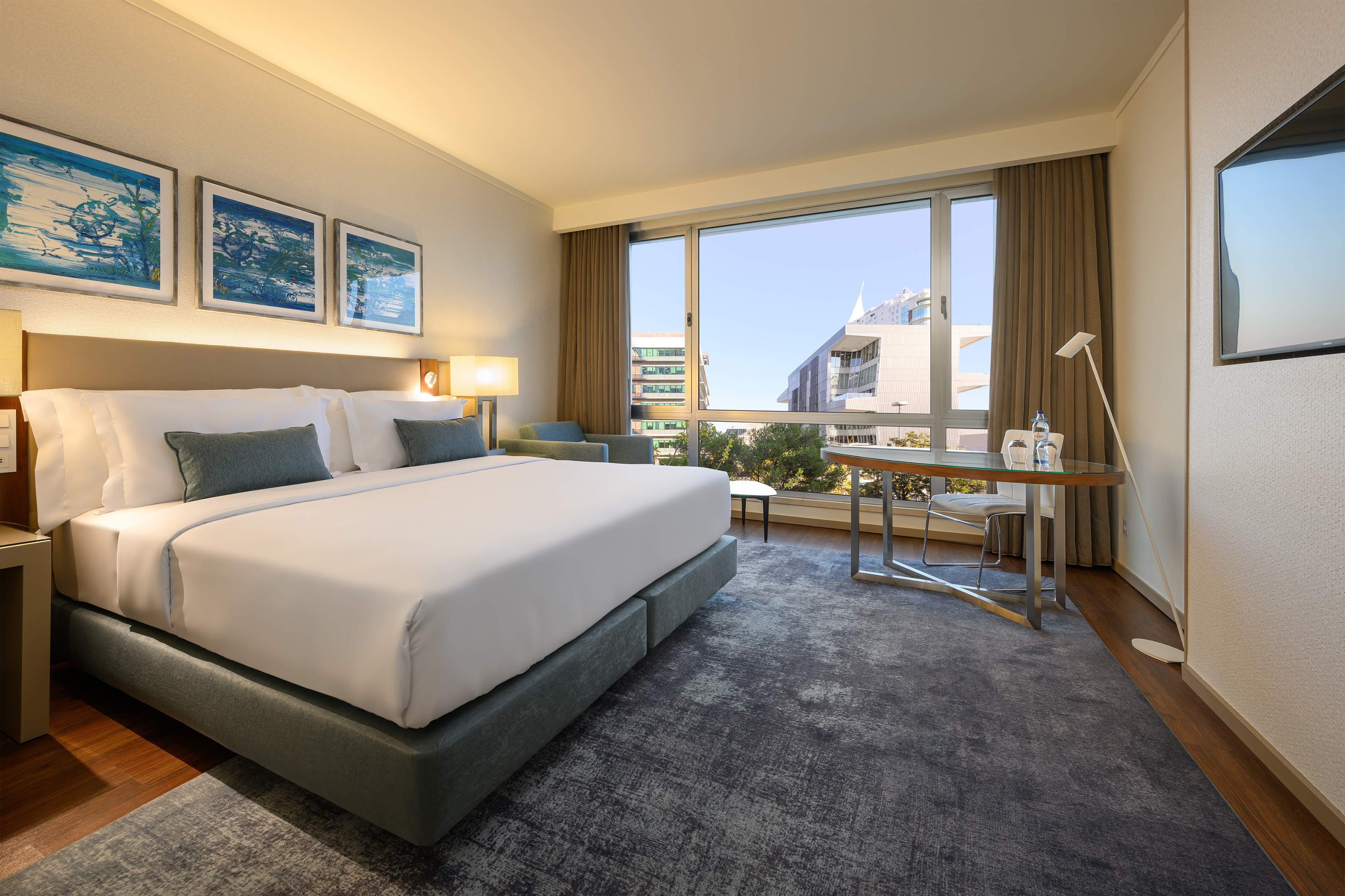 Modern hotel room with king bed, city view window, blue art, and desk.