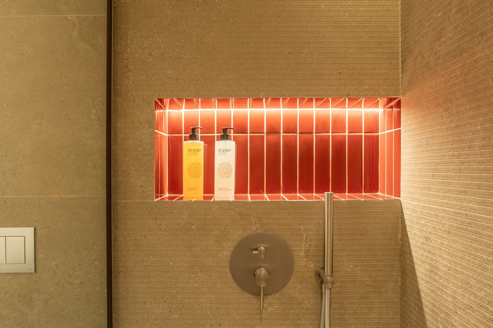 Modern shower with backlit red tile niche, argan products, and textured beige walls.