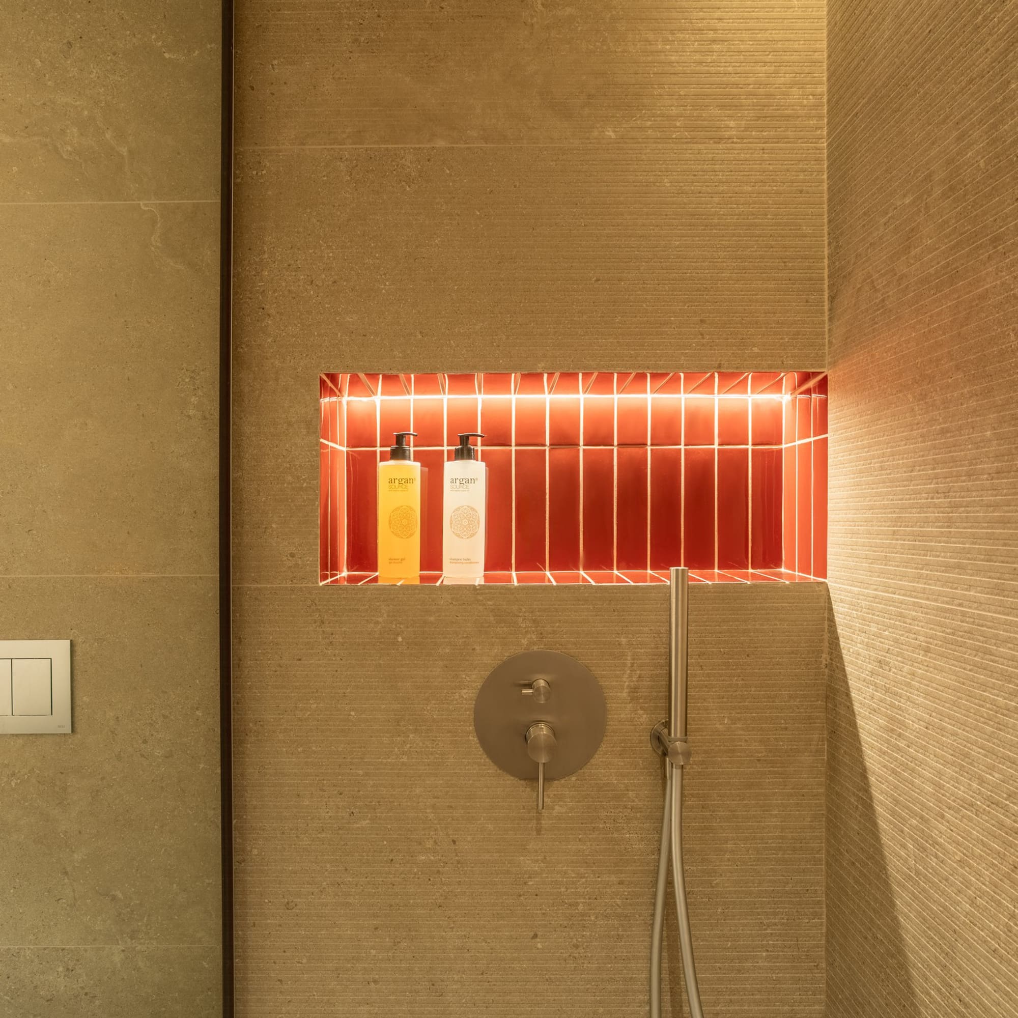 Modern shower with backlit red tile niche, argan products, and textured beige walls.