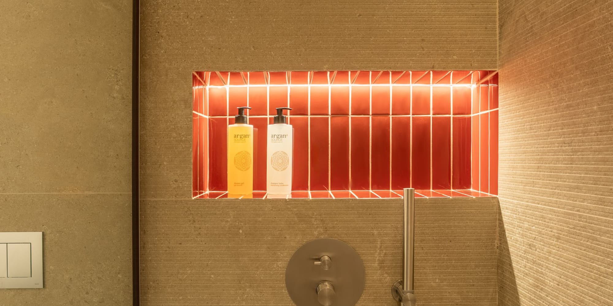 Modern shower with backlit red tile niche, argan products, and textured beige walls.